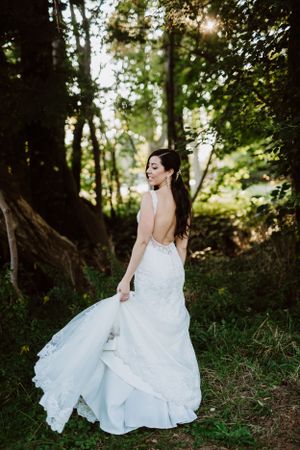 Elegant bride in backless lace wedding gown poses in sunlit forest setting.