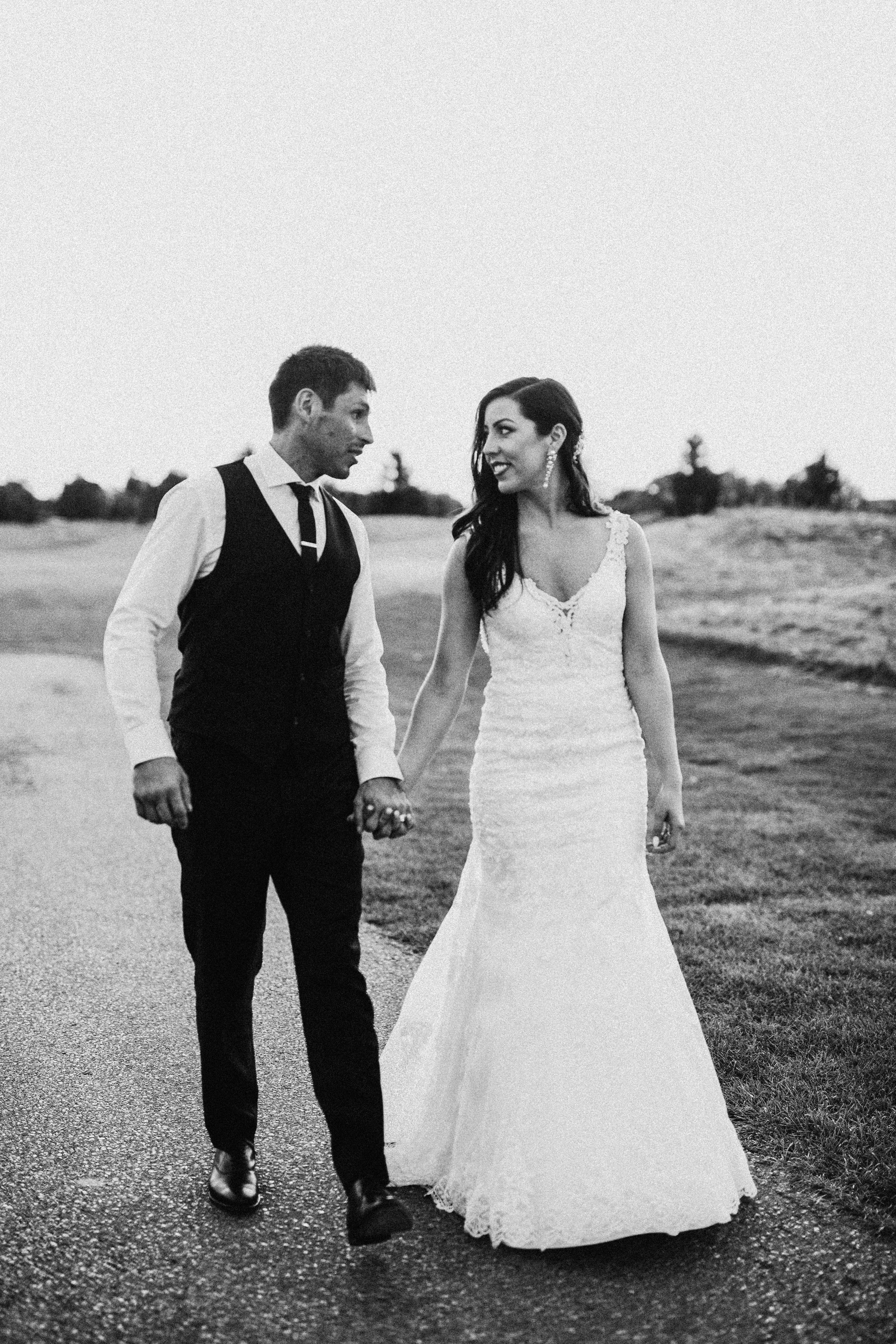 Black and white photo of newlyweds walking together on rural path in classic wedding attire.