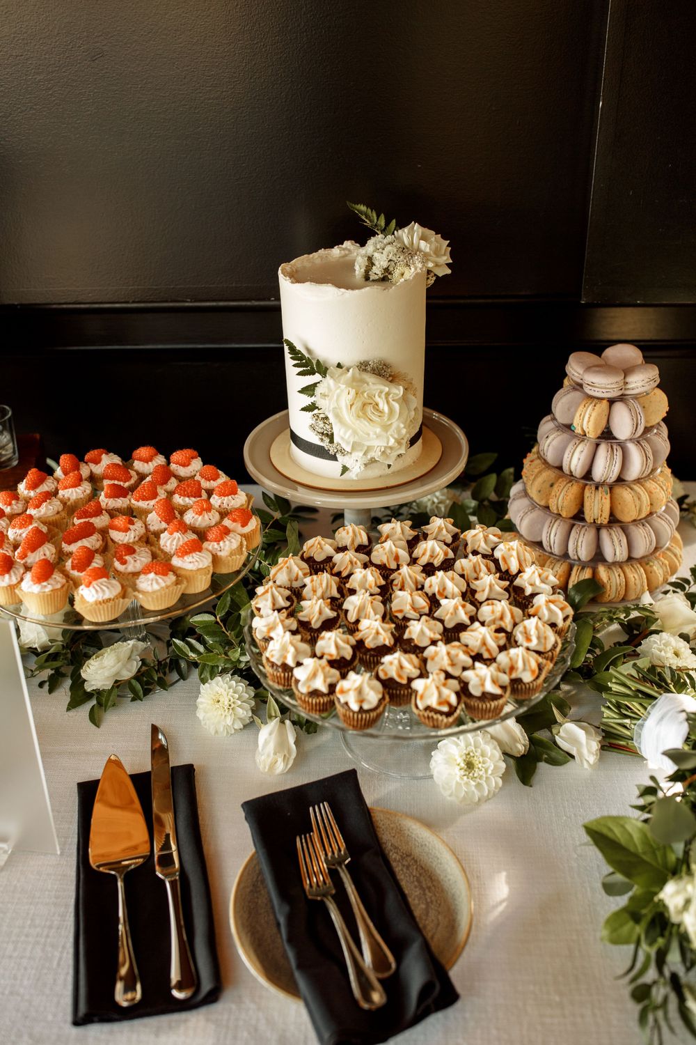 Elegant dessert display with wedding cake, macarons tower, cupcakes, and place settings decorated with white floral accents.