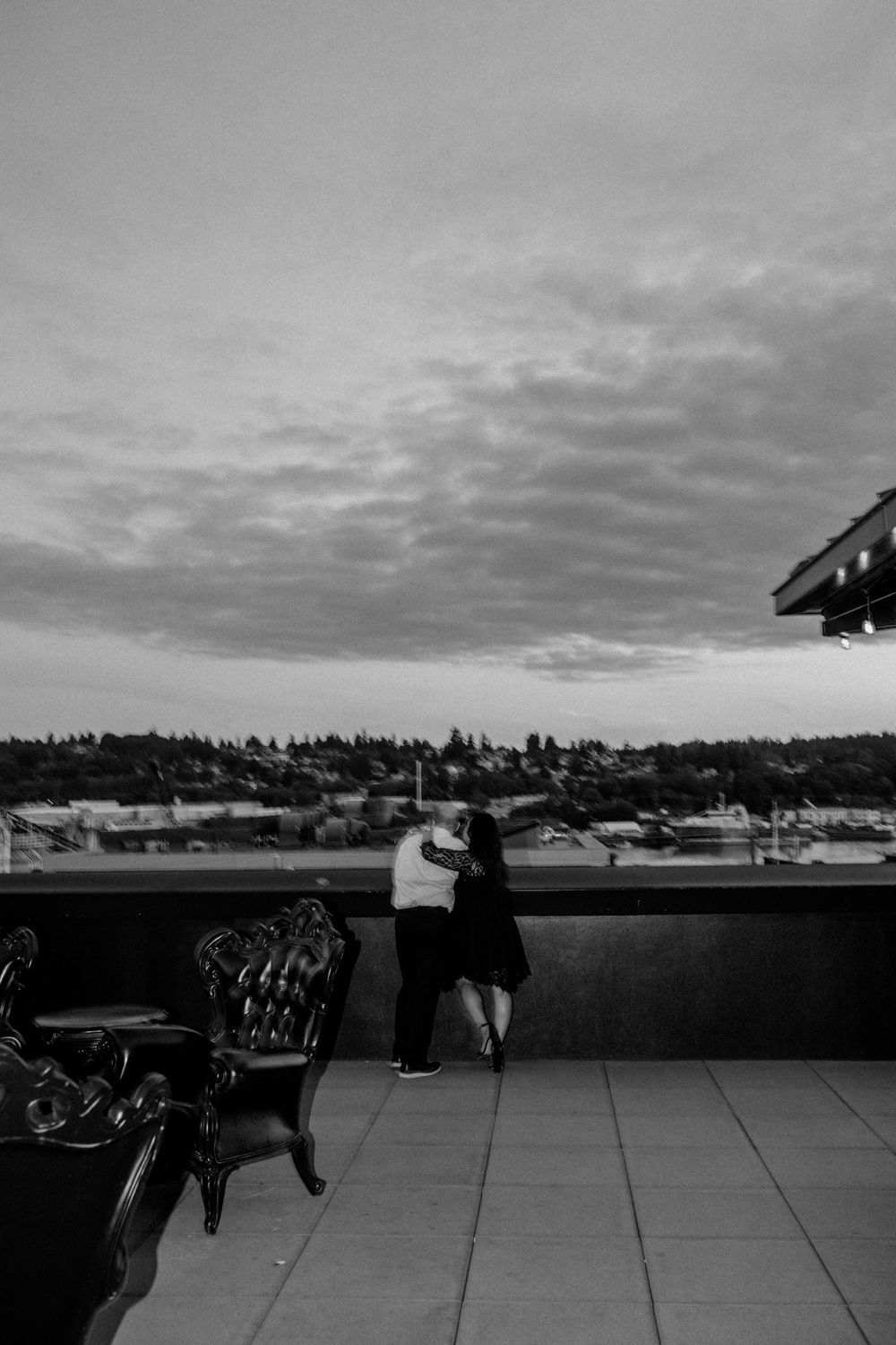 A couple embraces on a rooftop terrace overlooking a city skyline at dusk, with a decorative chair in the foreground.