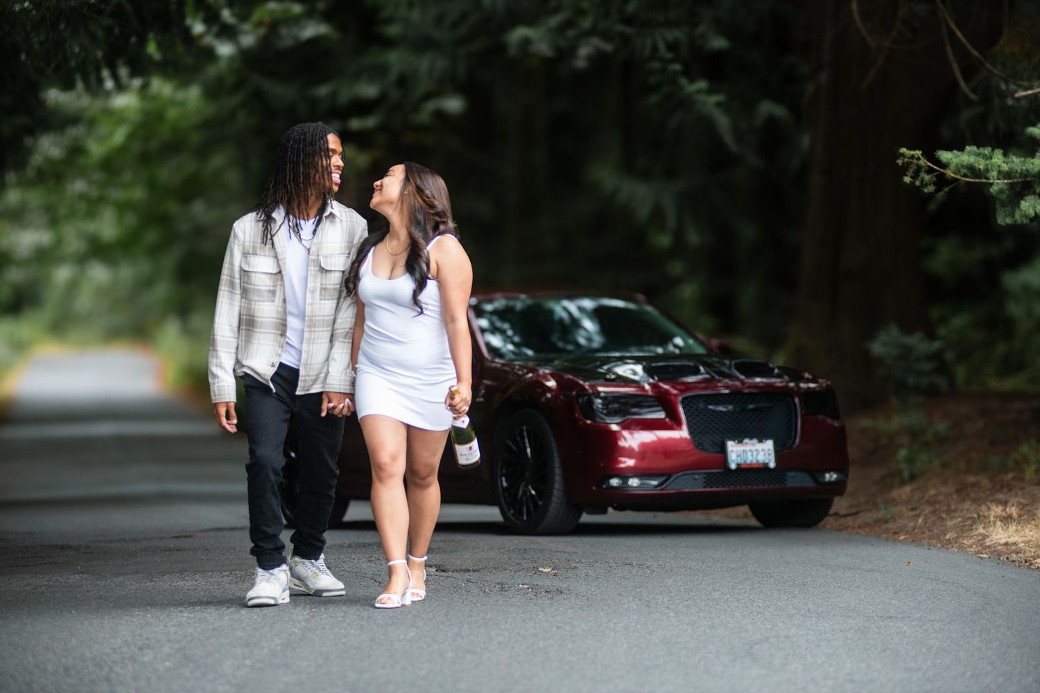 Couple walking on tree-lined road next to red sports car during engagement photoshoot.