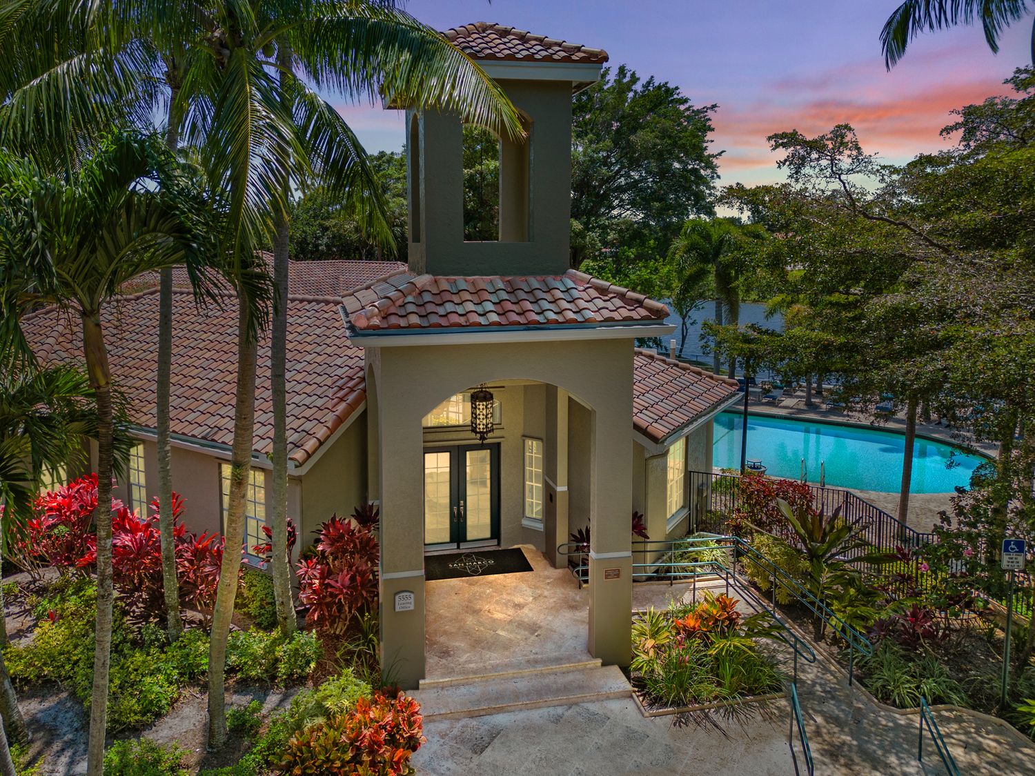 Luxurious Mediterranean villa with terracotta roof overlooks illuminated pool at twilight surrounded by tropical gardens and palms.