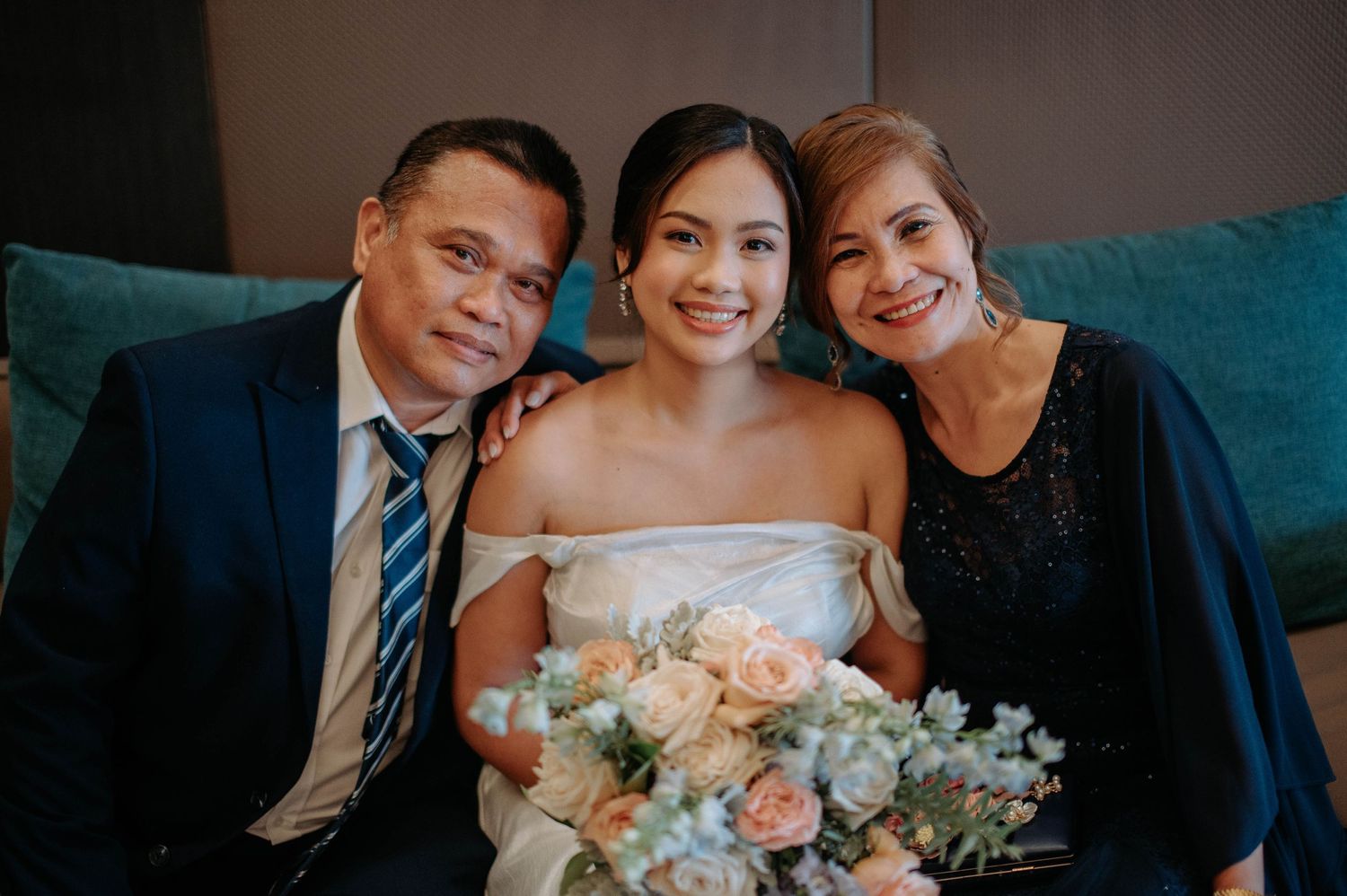 Wedding party portrait showing couple sitting on teal couch with bouquet of pink and white flowers.