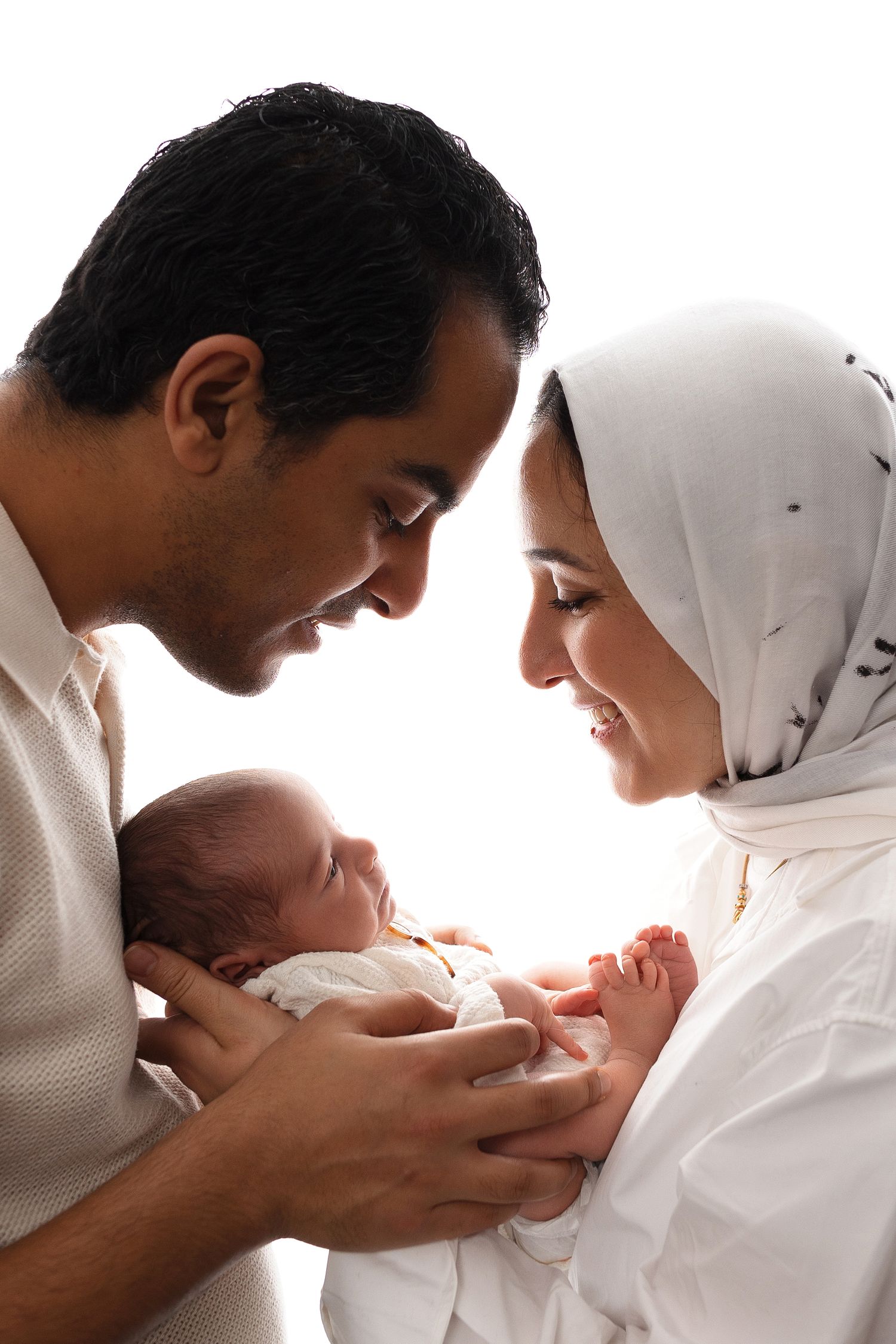 New parents in traditional Muslim attire lovingly hold and gaze at their newborn baby in intimate family portrait.
