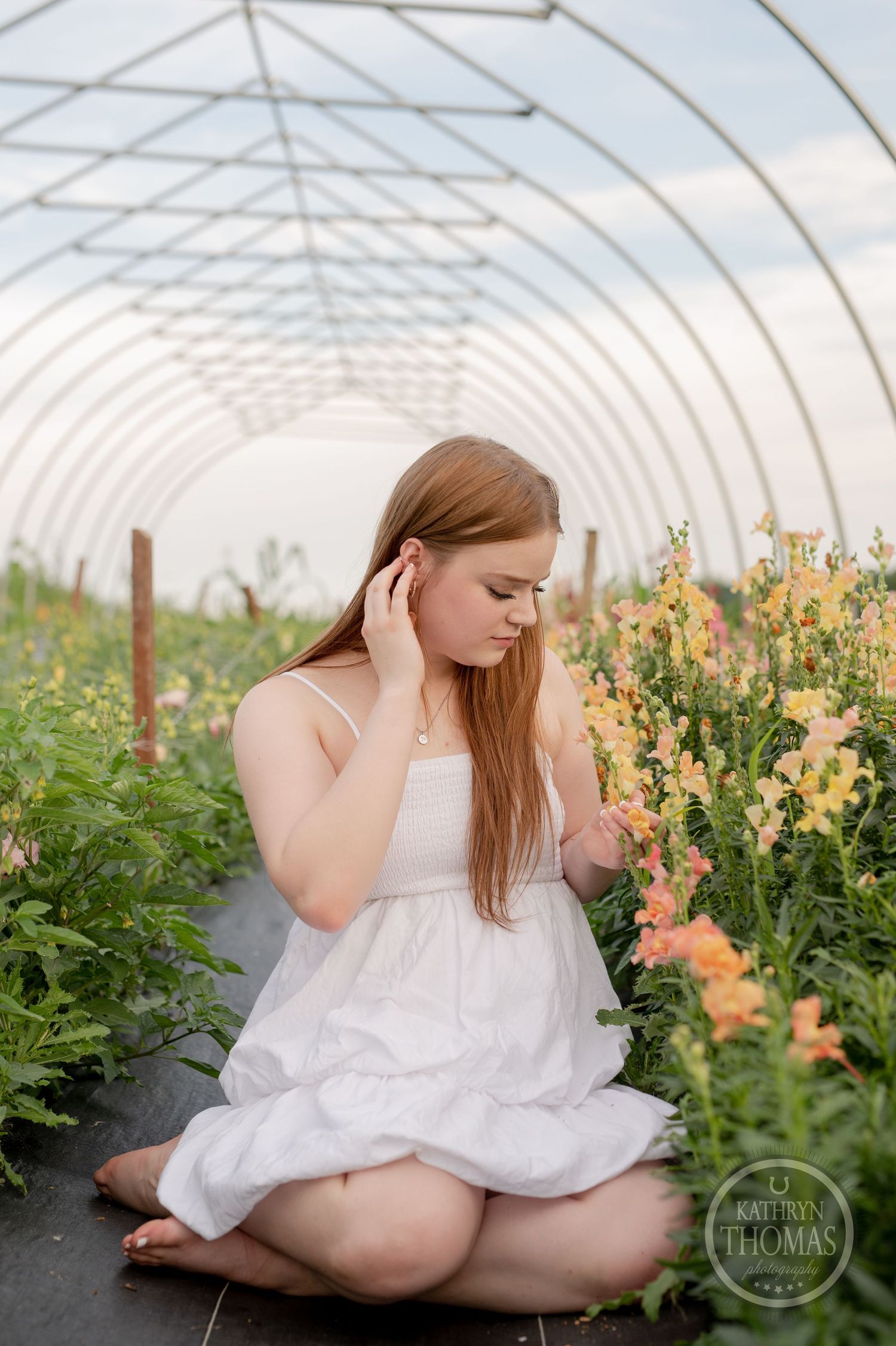 Portrait in white dress sitting inside greenhouse surrounded by blooming flowers.