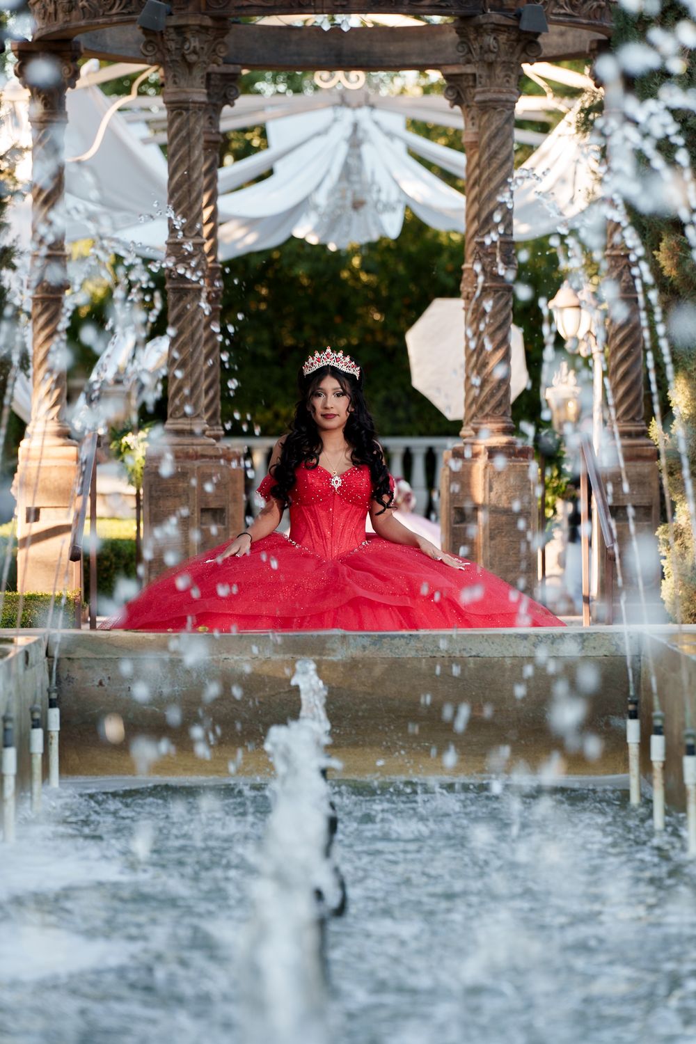 A stunning red quinceanera dress sparkles by a fountain in an elegant outdoor gazebo decorated with white fabric and flowers.