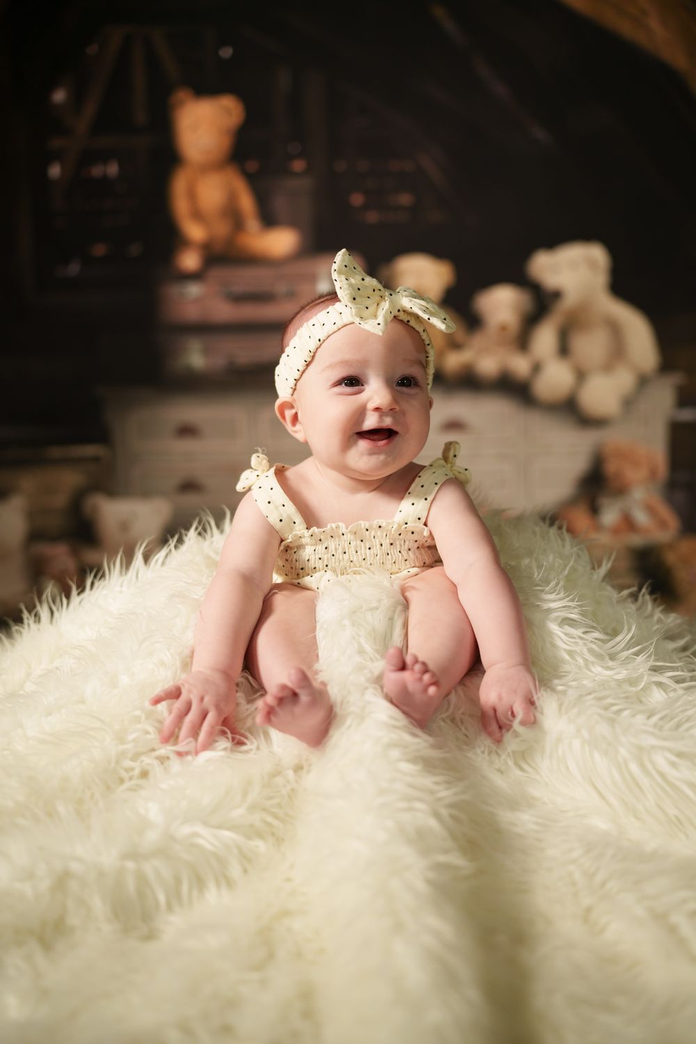 Baby wearing gold bow headband and dress sits on white fluffy rug with teddy bears in background.