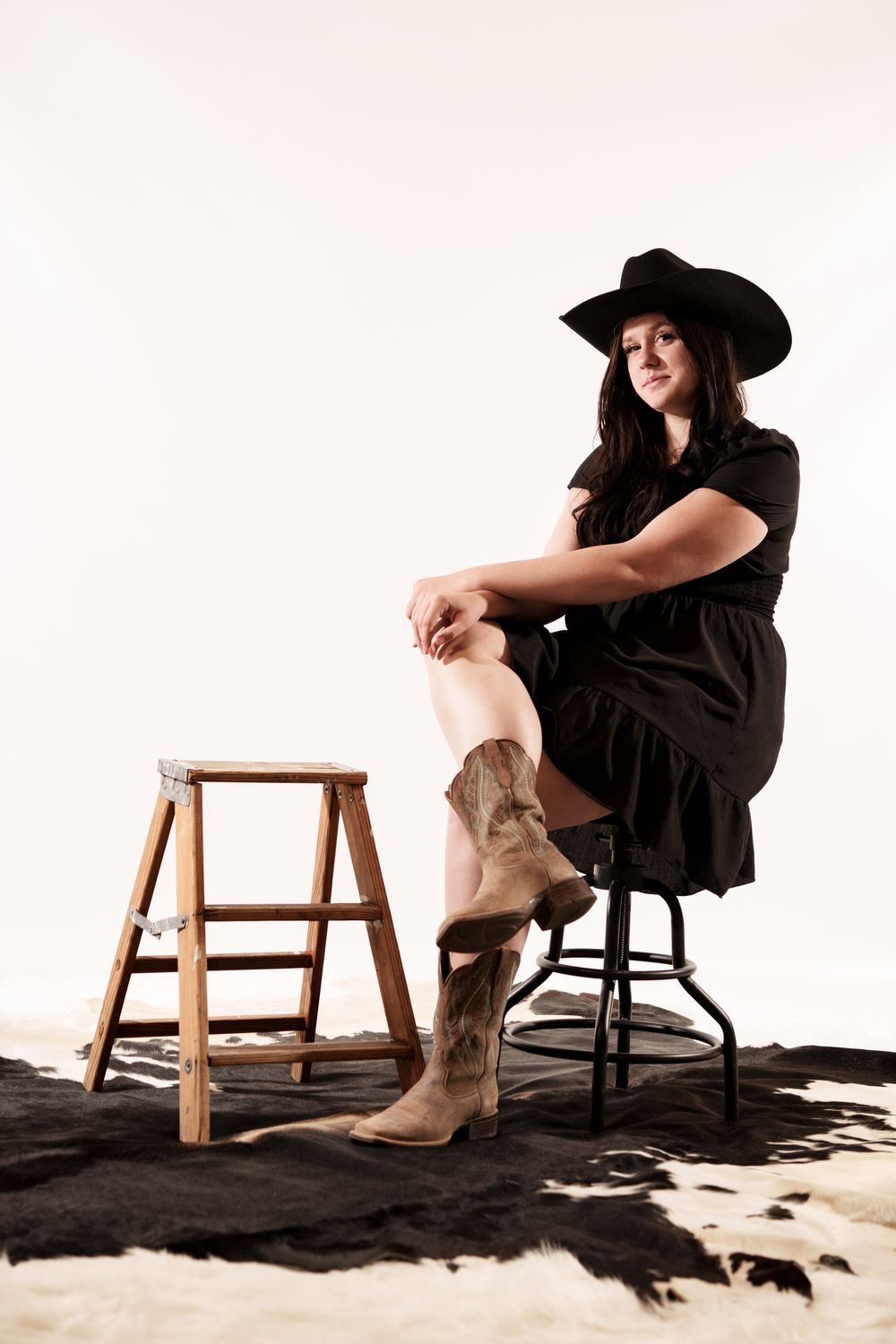 A rustic western-styled photo with cowboy boots, black dress, and wide-brimmed hat against a light backdrop.