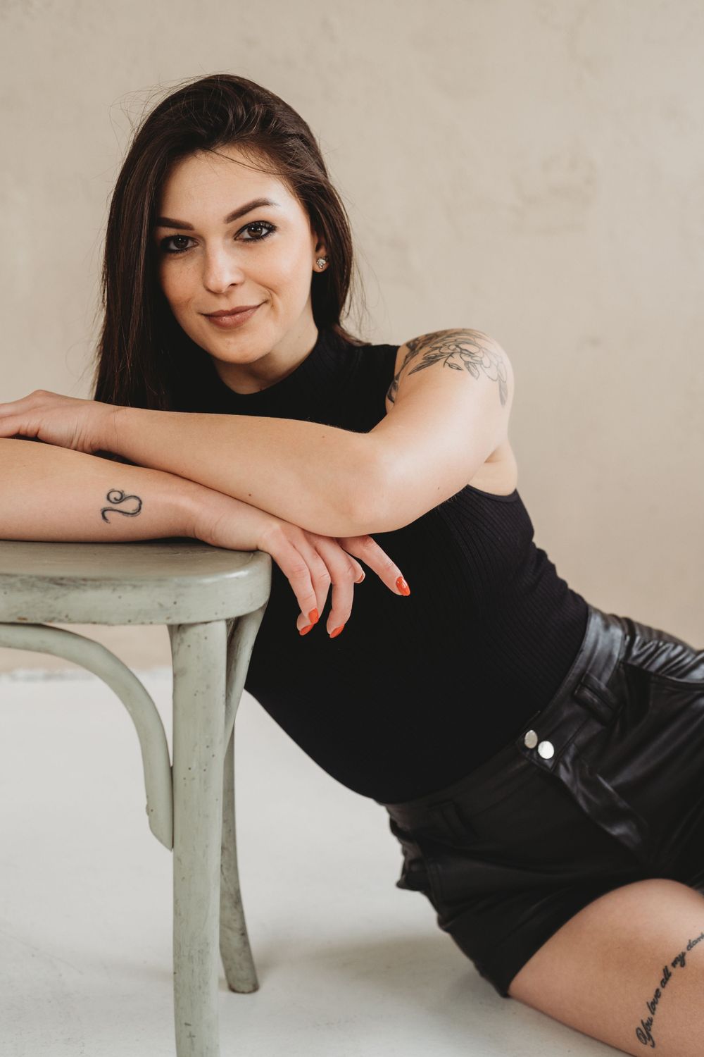 A person in a black sleeveless top and shorts poses casually against a mint green chair in a minimalist setting.