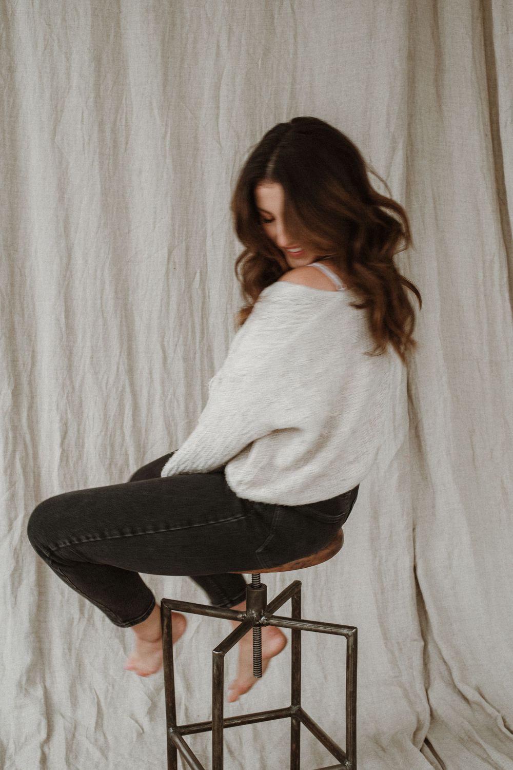 Woman in white sweater and dark jeans sits casually on industrial metal stool against light curtain backdrop.
