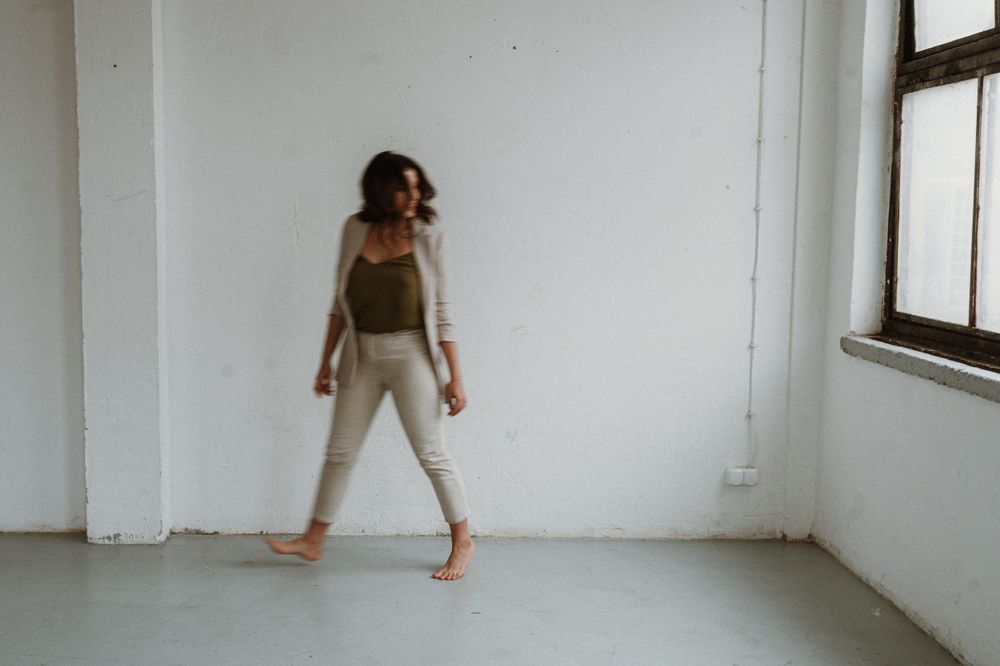 Someone stands barefoot in a minimalist white room wearing casual athletic wear and striking a dynamic pose.