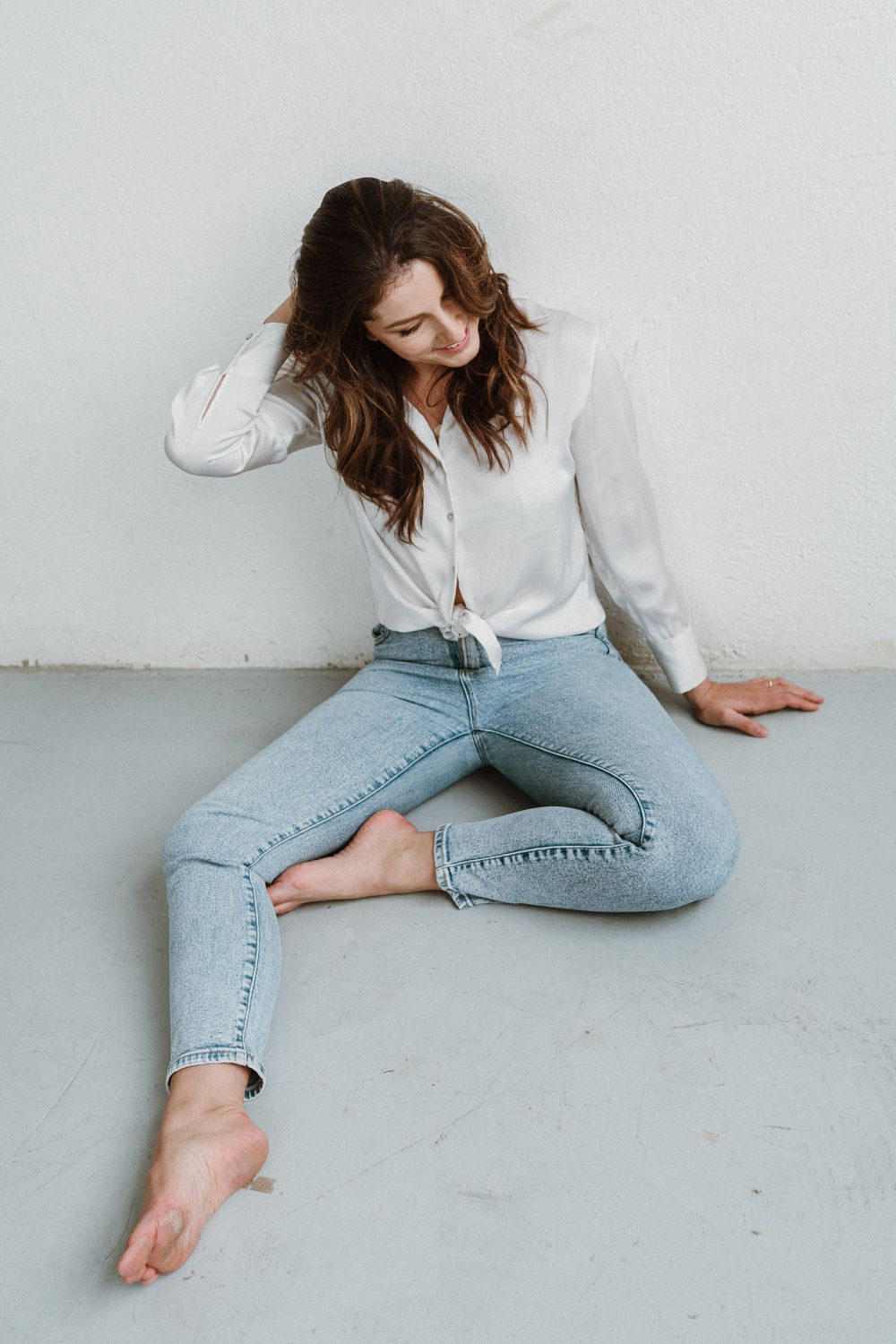 Model in white button down shirt and light wash jeans poses casually on concrete floor in minimalist fashion shoot.