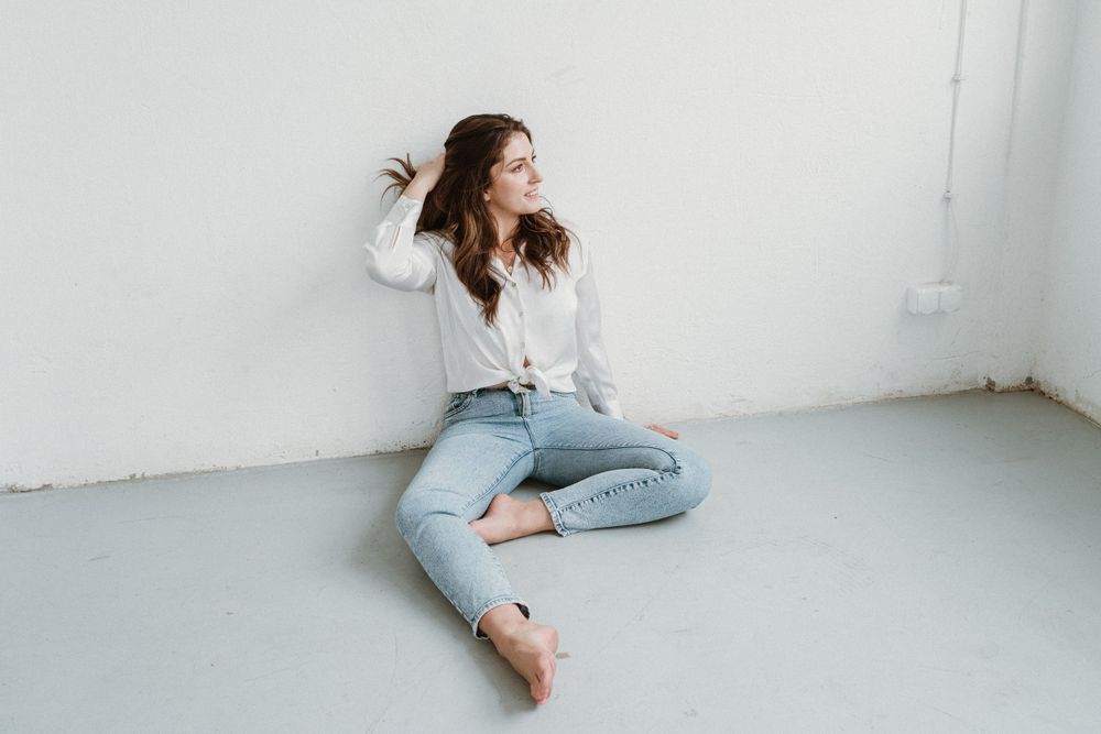 Person in white sweater and ripped jeans sitting against white wall with hair in motion.