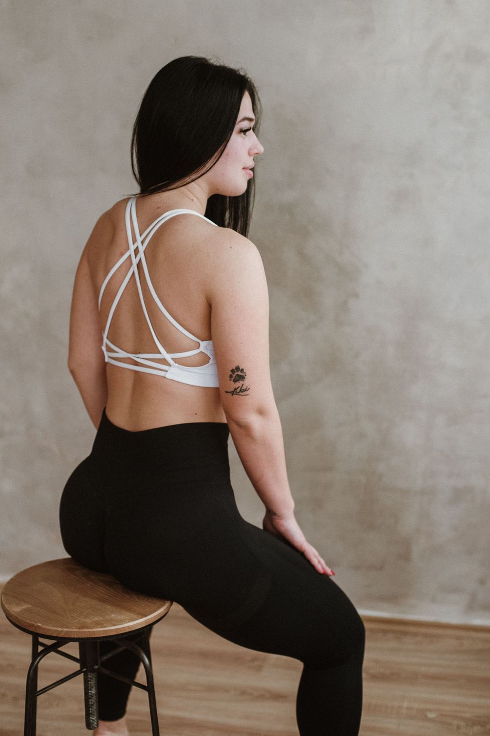 Profile portrait of someone wearing a white strappy sports bra and black pants while sitting on a wooden stool.
