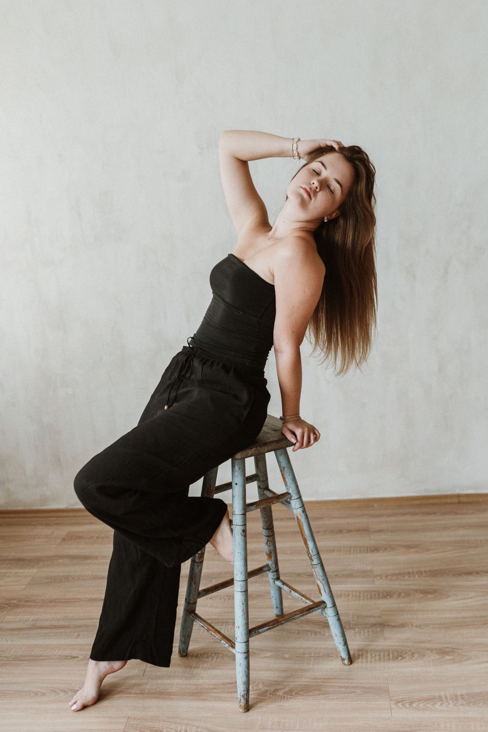 Woman in black jumpsuit poses expressively while sitting on a blue industrial stool against white wall.