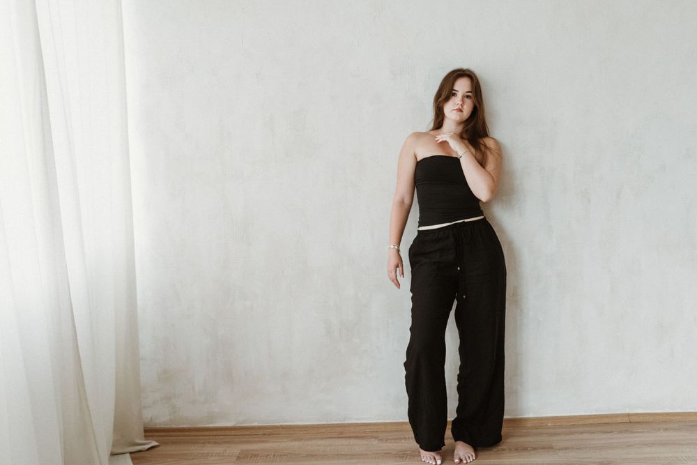Modern minimalist portrait against white wall showing black sleeveless top and matching pants outfit.