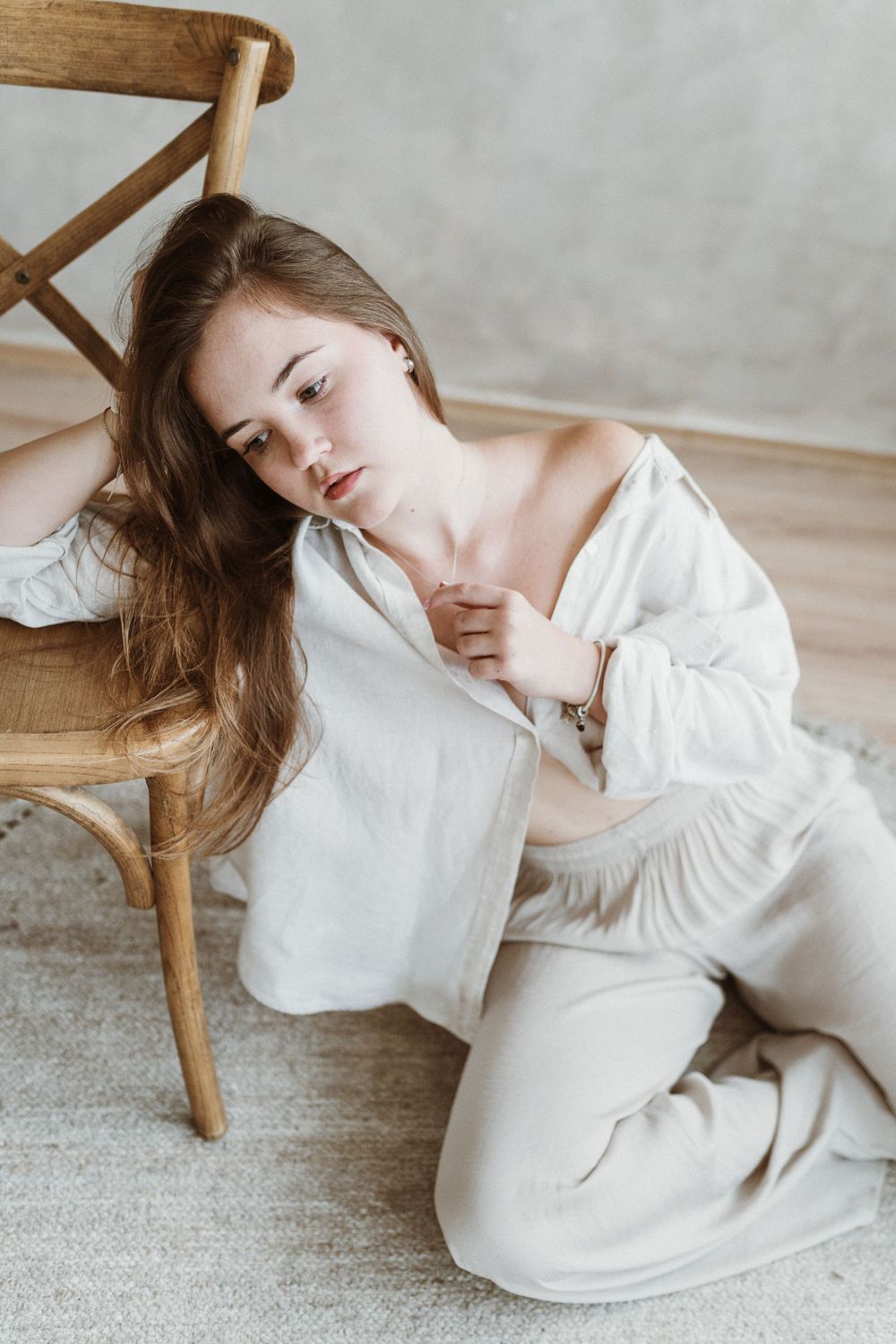 Person in white loungewear rests against wooden chair on light carpet in minimalist setting.