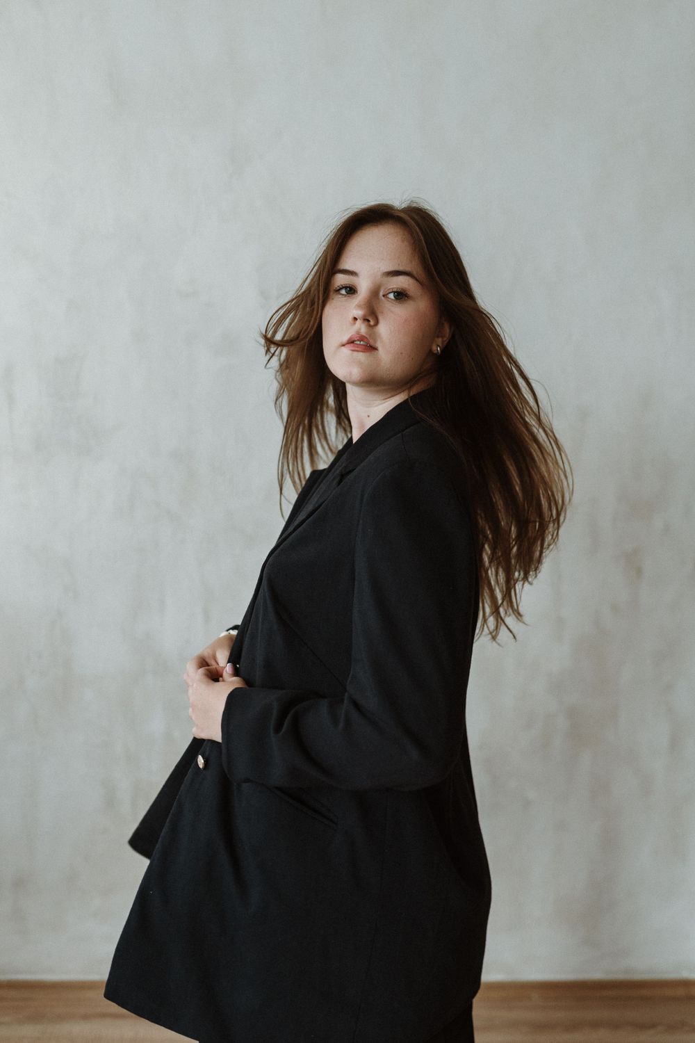 A figure in a black blazer poses dramatically against a light gray wall with flowing brown hair in motion.