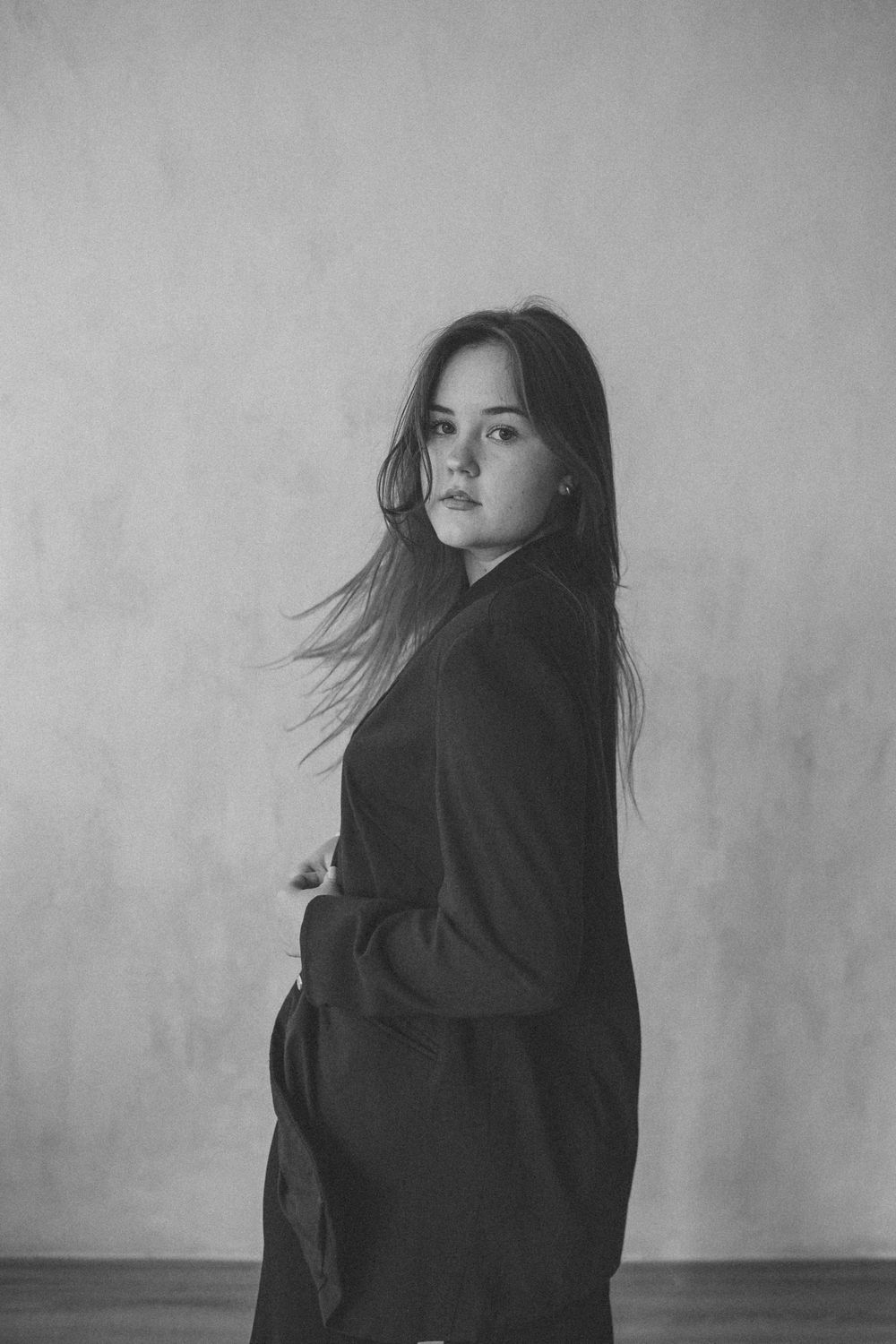Black and white portrait of person in dark coat with flowing hair against minimalist background.