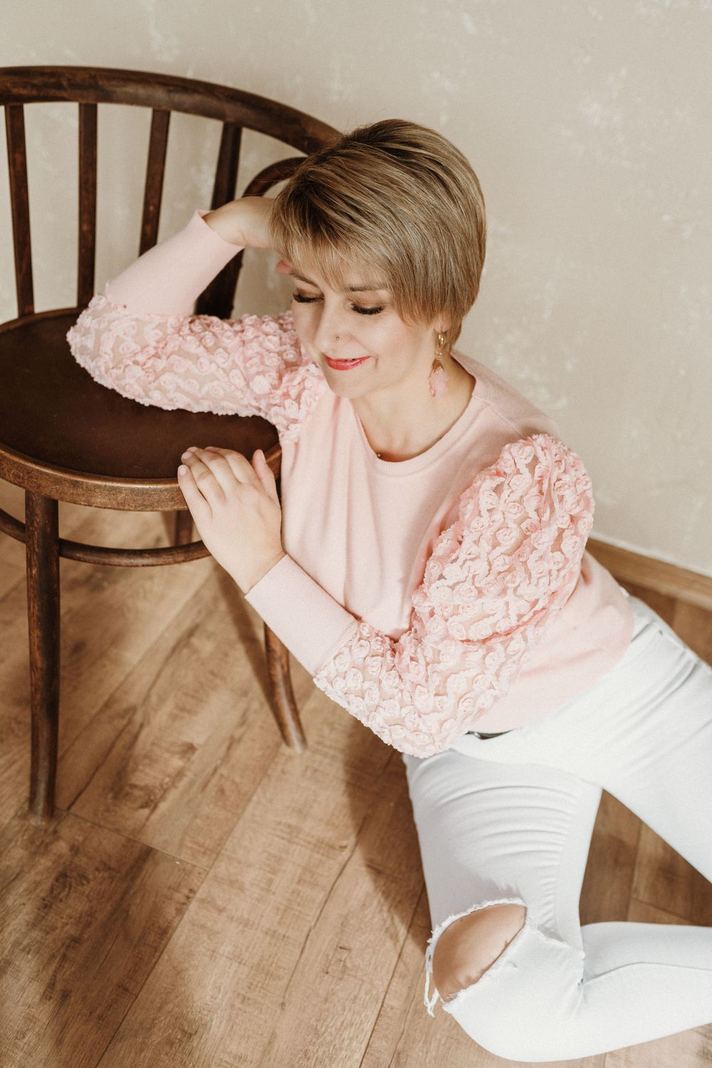 A feminine figure in a cozy pink sweater with textured sleeves and white ripped jeans poses on a wooden chair.