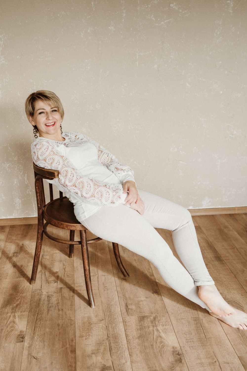 Someone relaxes in an elegant white lace outfit while lounging on a wooden chair against a neutral backdrop.