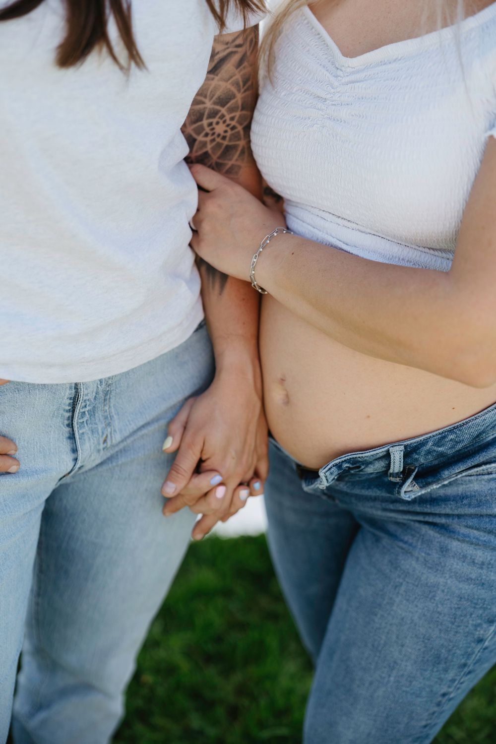 Two women in white tops and jeans hold hands outdoors, one visibly pregnant with a tattoo on her arm.