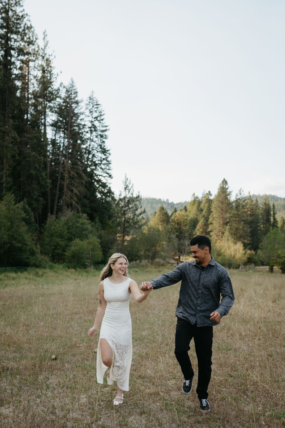 A joyful couple holds hands while walking through a scenic meadow surrounded by tall pine trees.