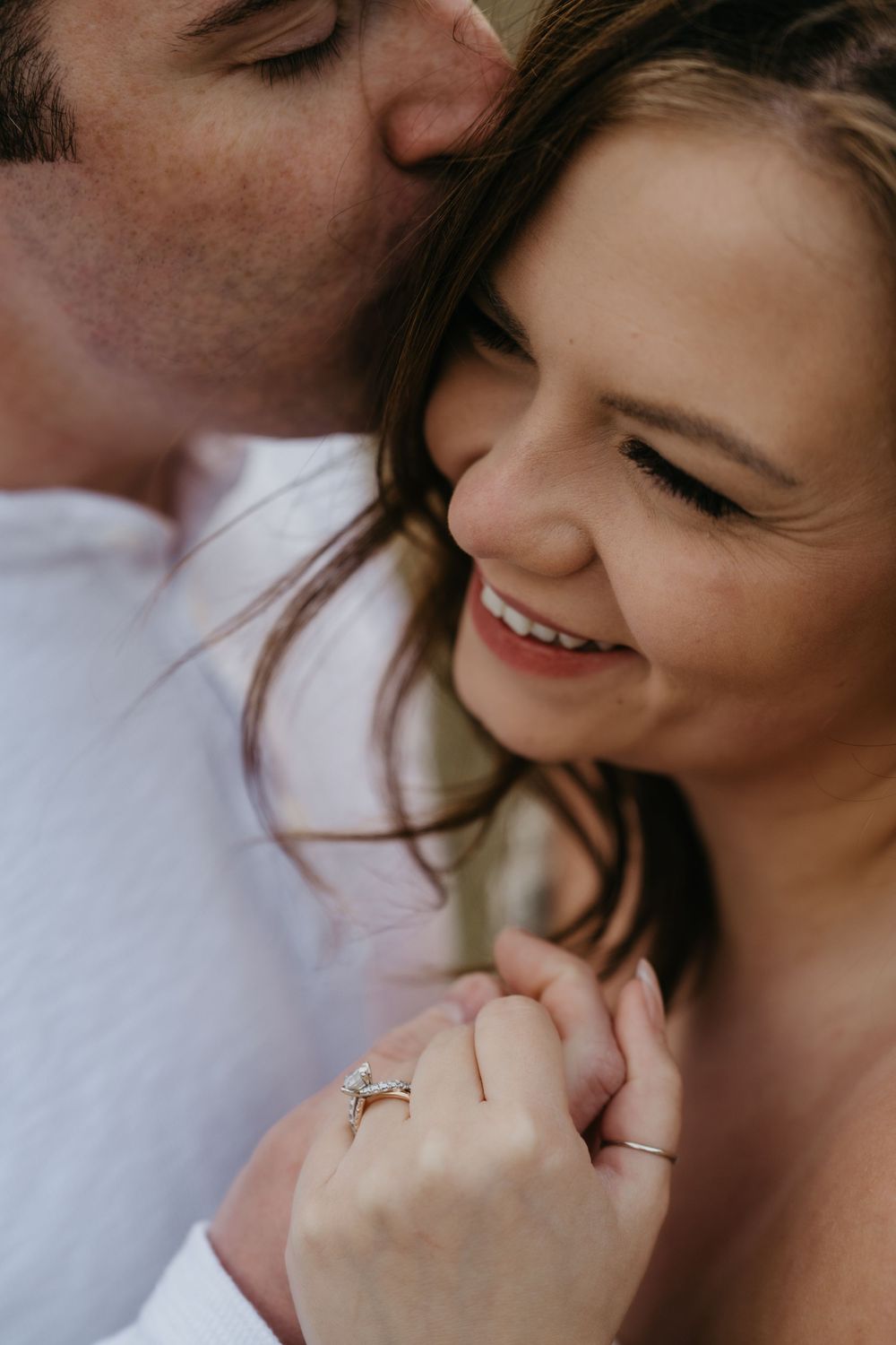 A smiling woman with an engagement ring receives a tender kiss on the cheek from her partner during a romantic couples photo session.