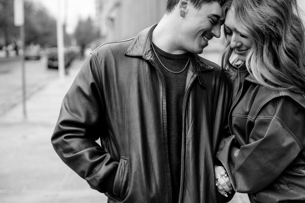 Black and white intimate moment between couple sharing laughter on city sidewalk in leather jackets.