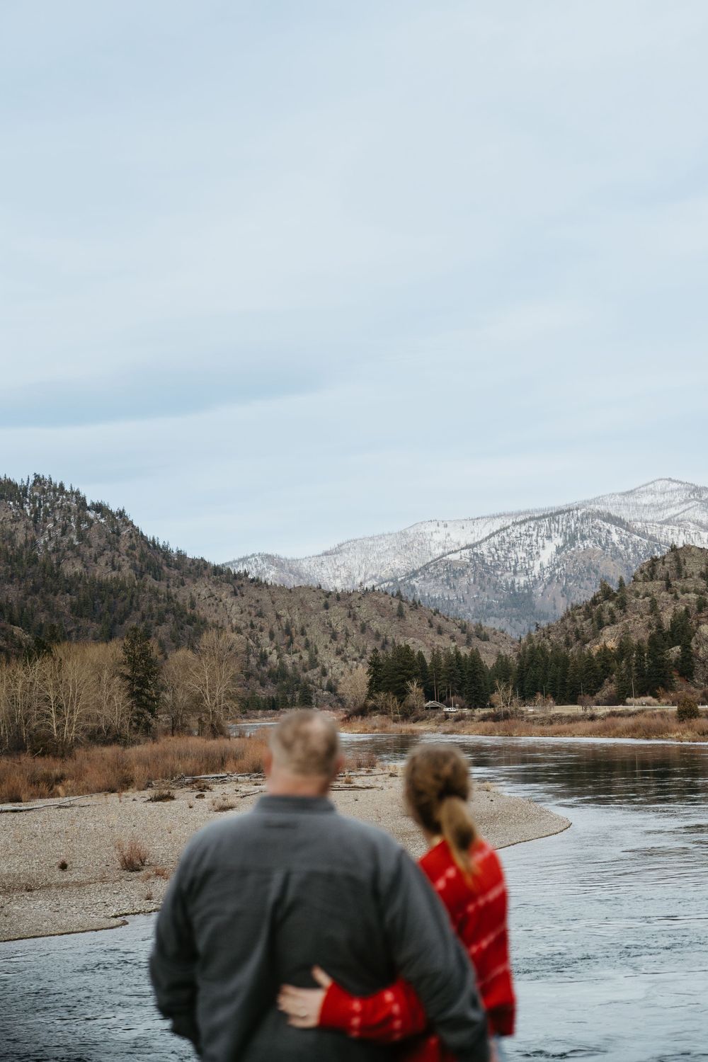 A couple stands at the edge of a winding river with snow-capped mountains in the background on a cloudy winter day.