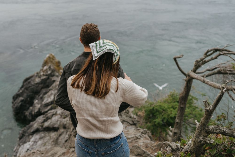 A photographer wearing a sweater and jeans stands on a coastal cliff photographing a seagull over misty waters.