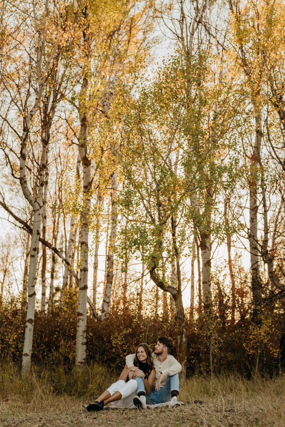 Couple relaxing under tall autumn birch trees in golden evening light.
