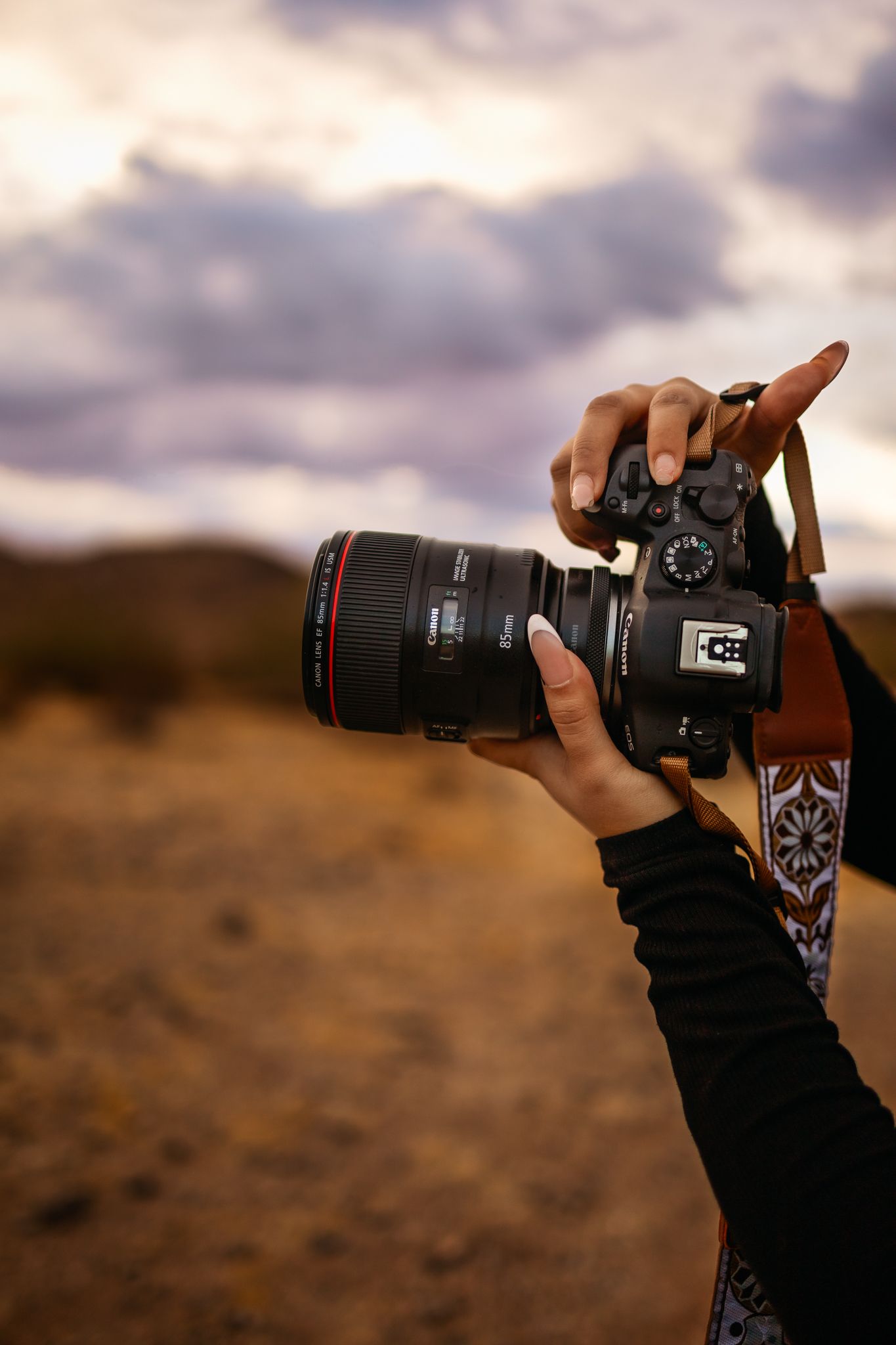 Close-up shot of hands holding a professional photography camera with telephoto lens against a dramatic sunset sky.