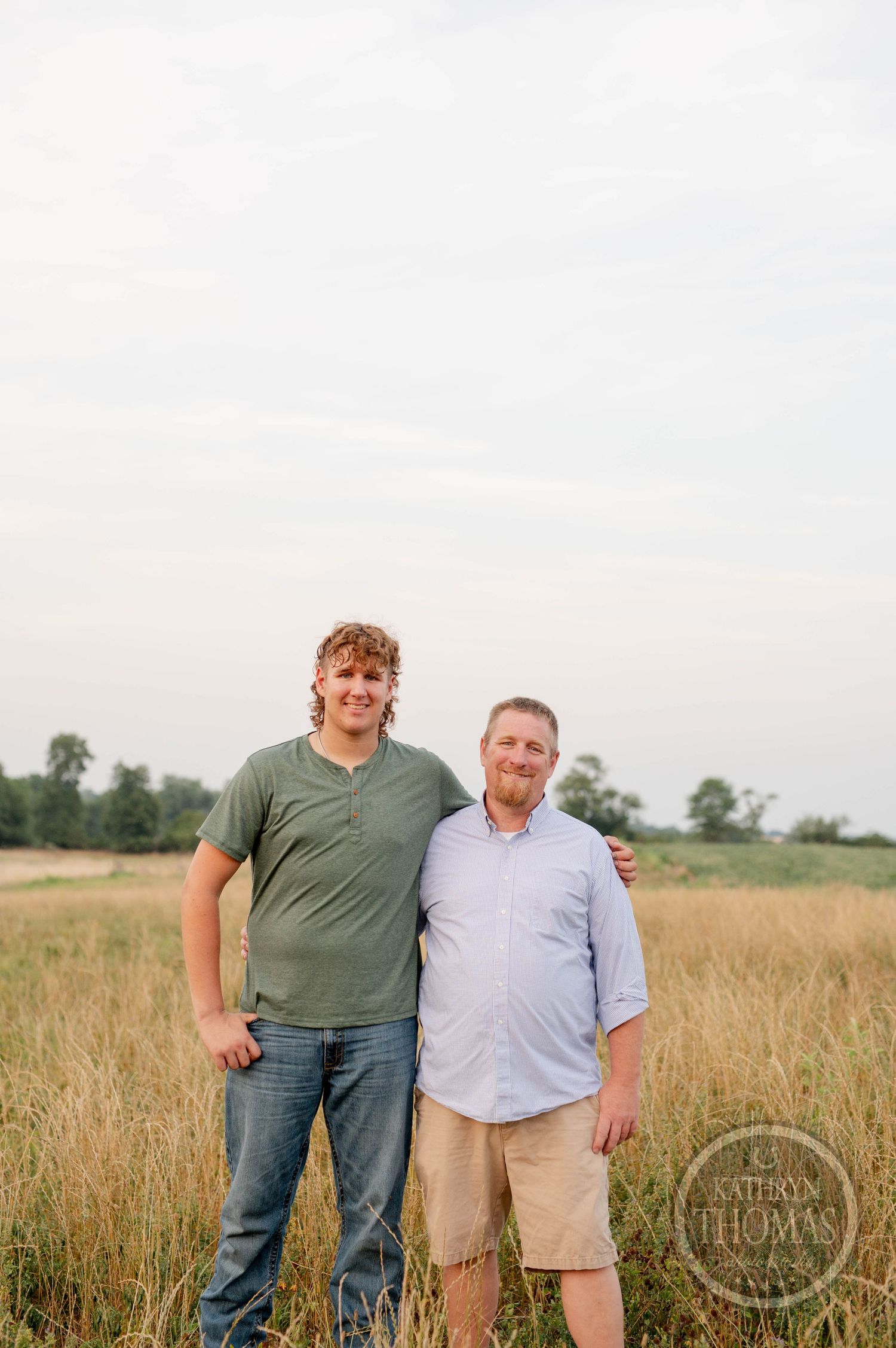Two people stand together in a grassy field during golden hour, wearing casual summer attire.