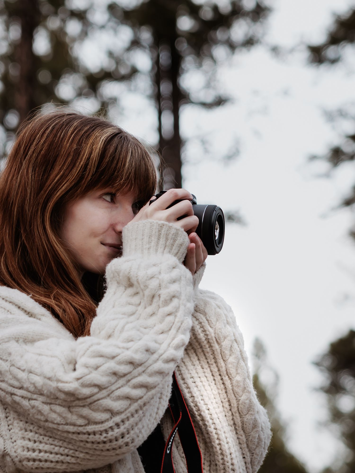 Photographer in cream cable knit sweater takes photos with camera against forest backdrop.