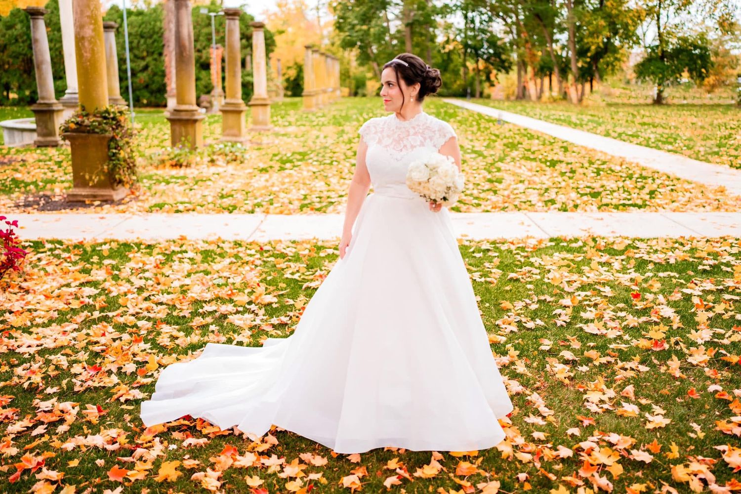 A bride in a white wedding gown stands in a garden surrounded by autumn leaves and stone pillars.