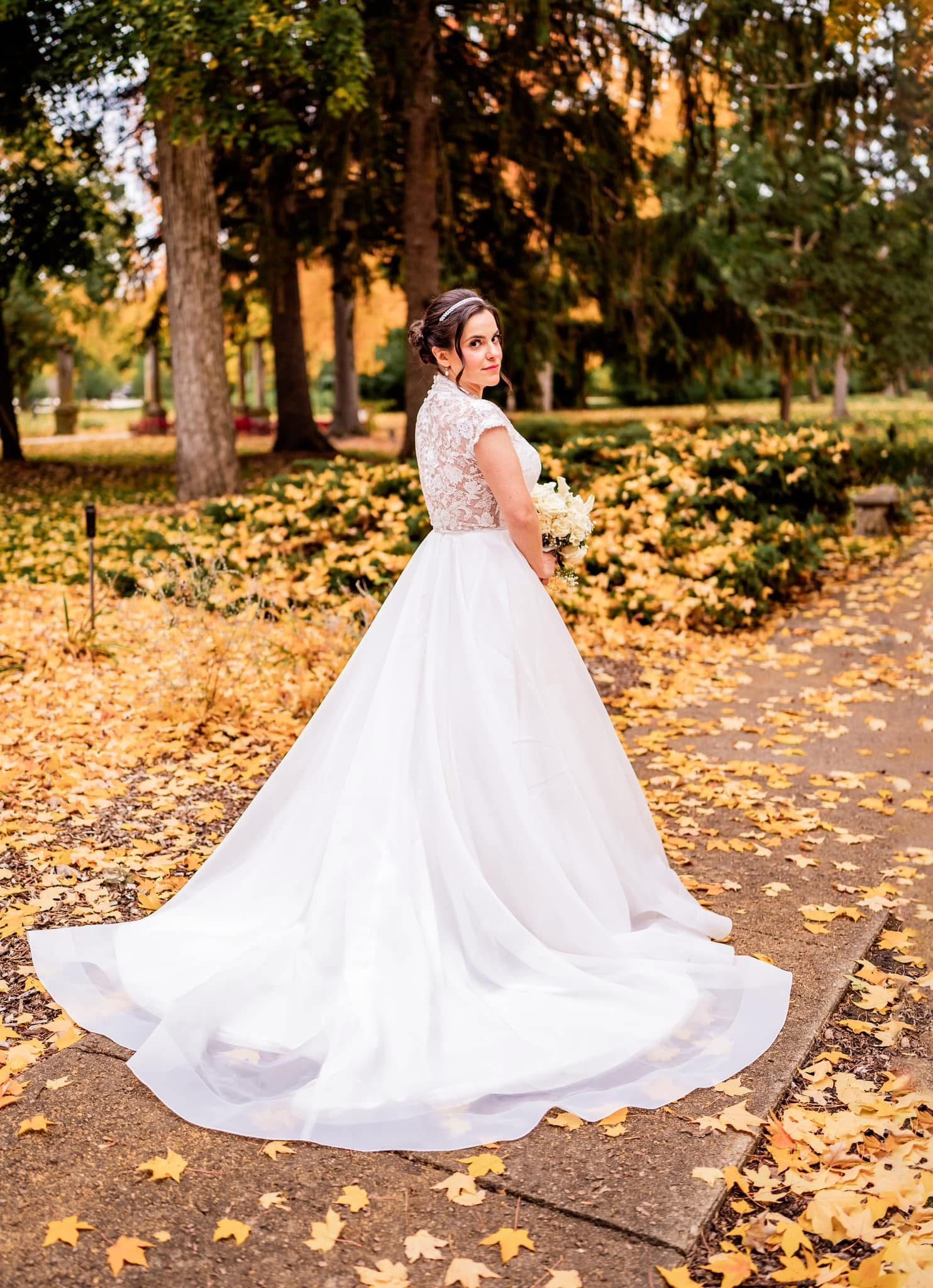 A stunning white wedding dress with lace details twirls in an autumn setting with golden fallen leaves.