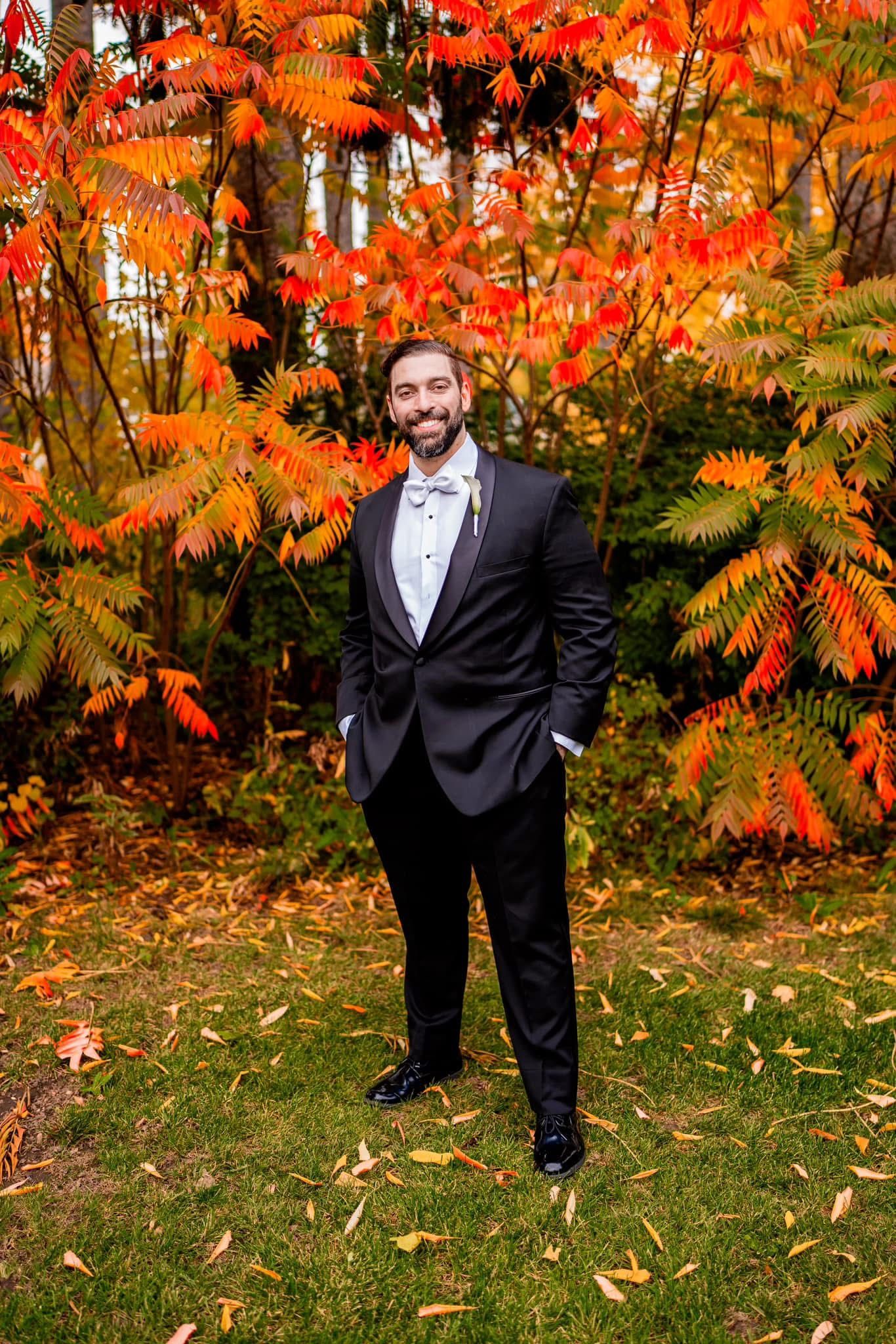 A groom in a black tuxedo poses against vibrant orange autumn foliage in a garden setting.