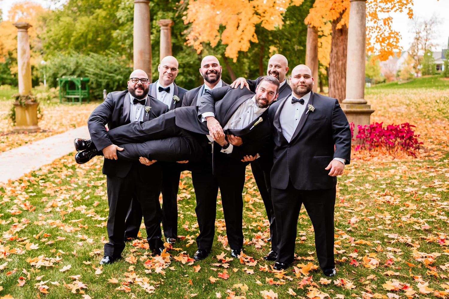 Groomsmen in black suits playfully lift one member of their group in a beautiful fall garden setting with autumn leaves.