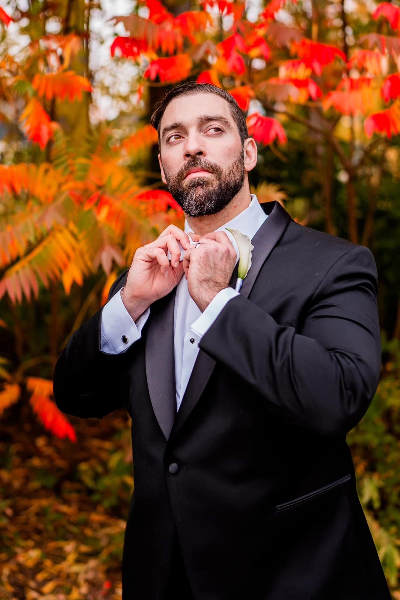 A formal wedding portrait against vibrant red autumn foliage while adjusting a bowtie.