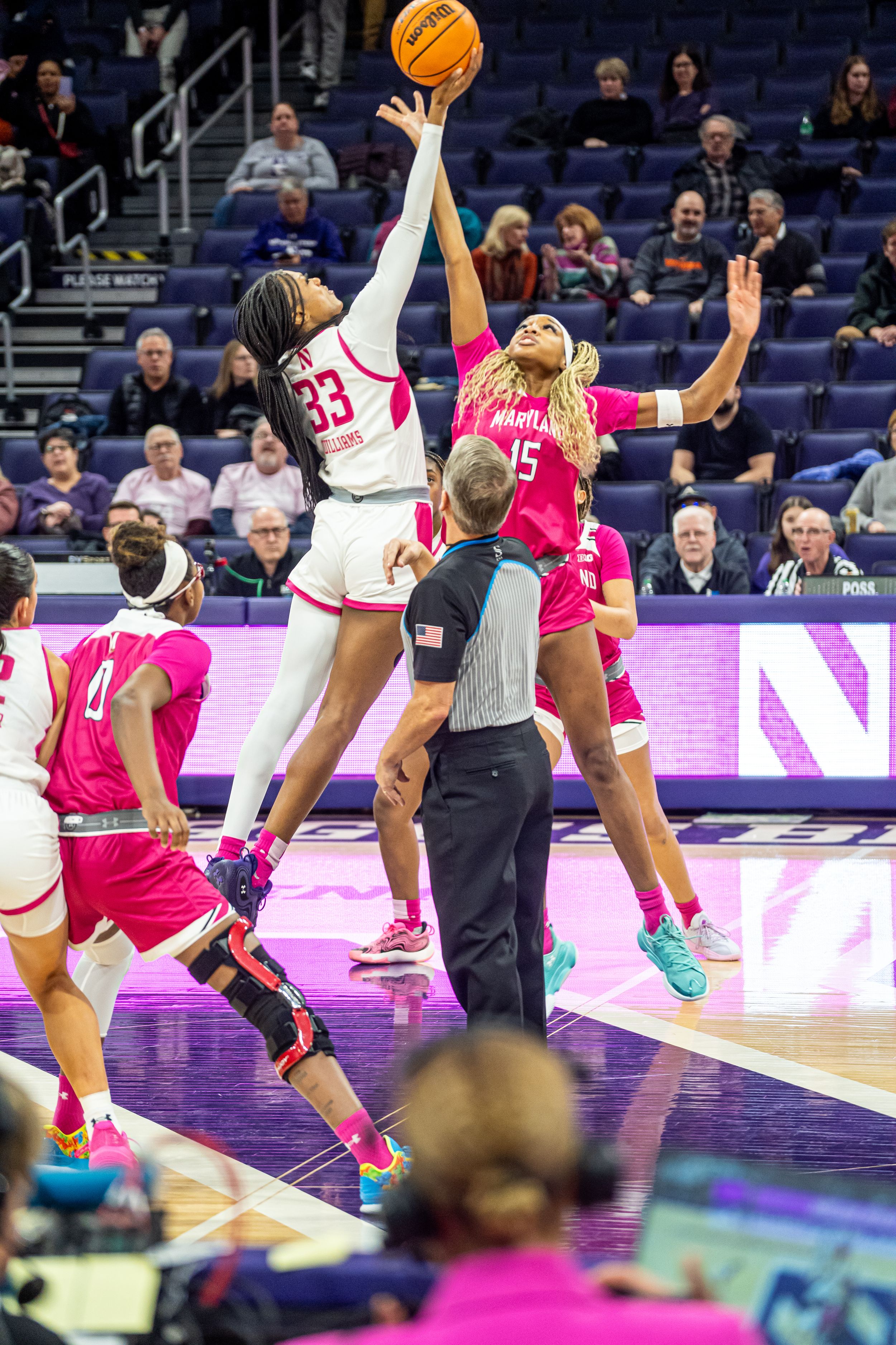 Basketball players in white and pink uniforms leap high to contest for the ball during an intense game moment.