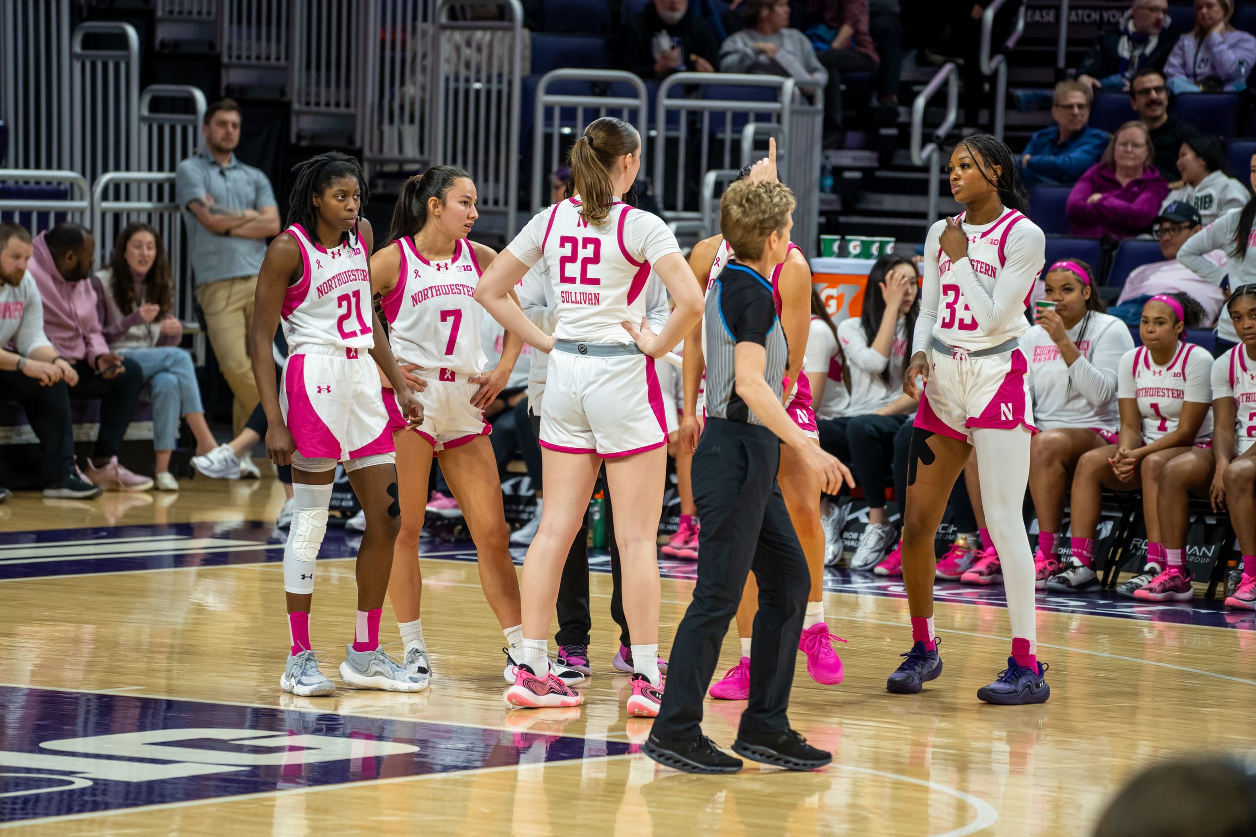 Basketball players and coach gather on the court during a game wearing pink and white uniforms.