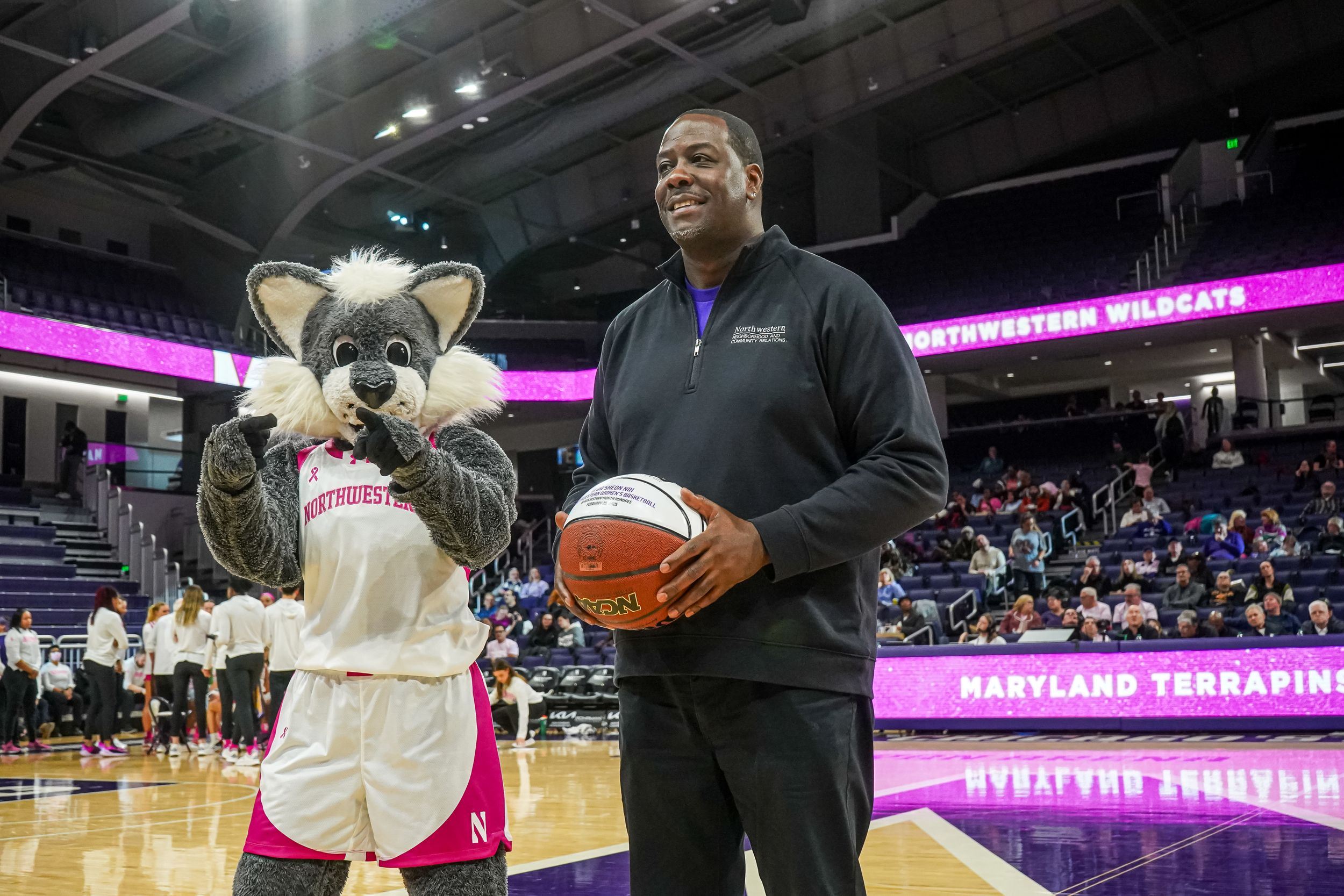 Basketball event on court features team mascot in white uniform and staff member holding game ball during timeout.