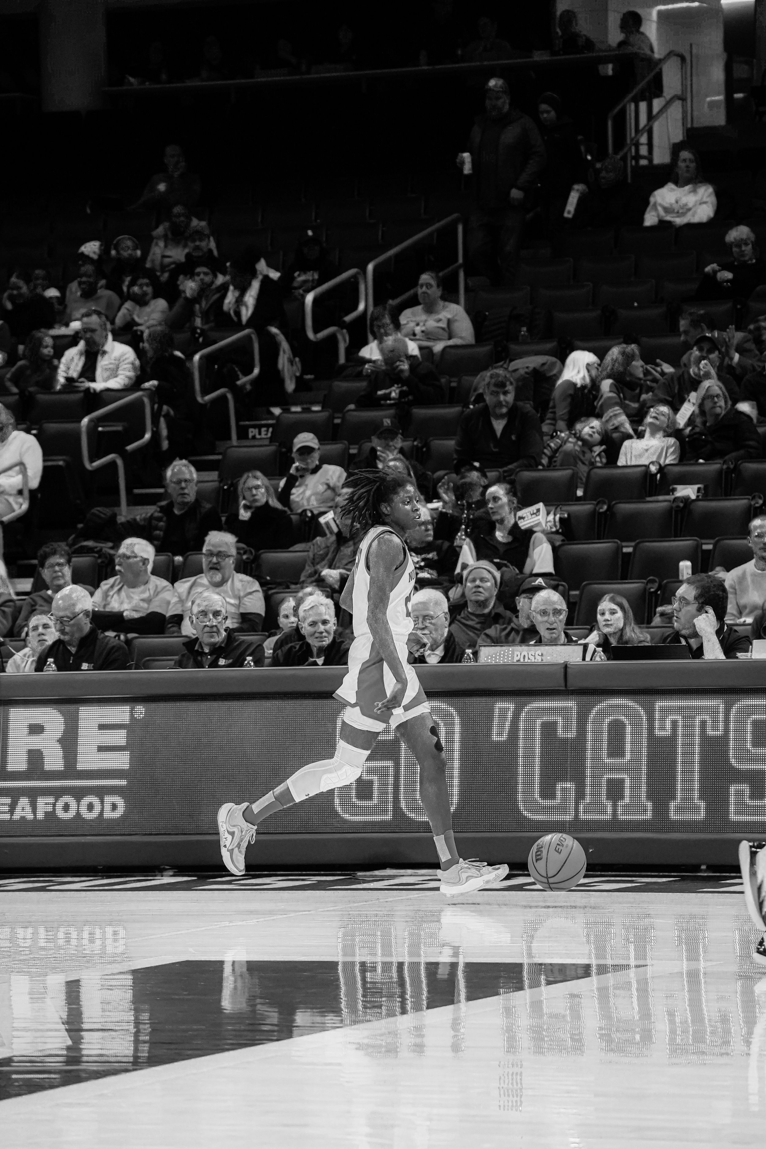 A basketball player dribbles down the court during a game with spectators watching from the stands.