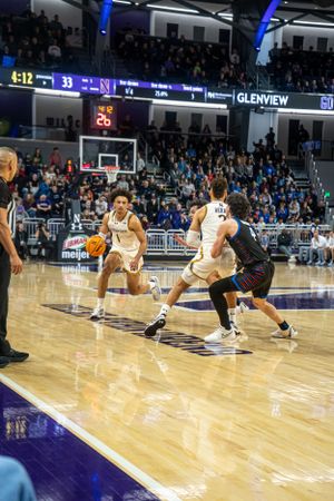 Basketball players compete on a polished court during an intense game with blue lighting and stadium seating visible.