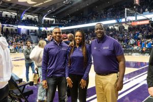 Three people wearing purple outfits stand together smiling at a basketball arena during an event.