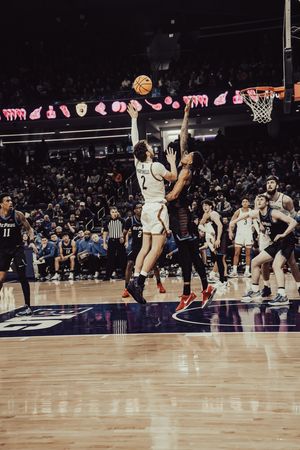 Basketball players leap for the ball during an intense game on a wooden court with stadium lights and pink decorations.
