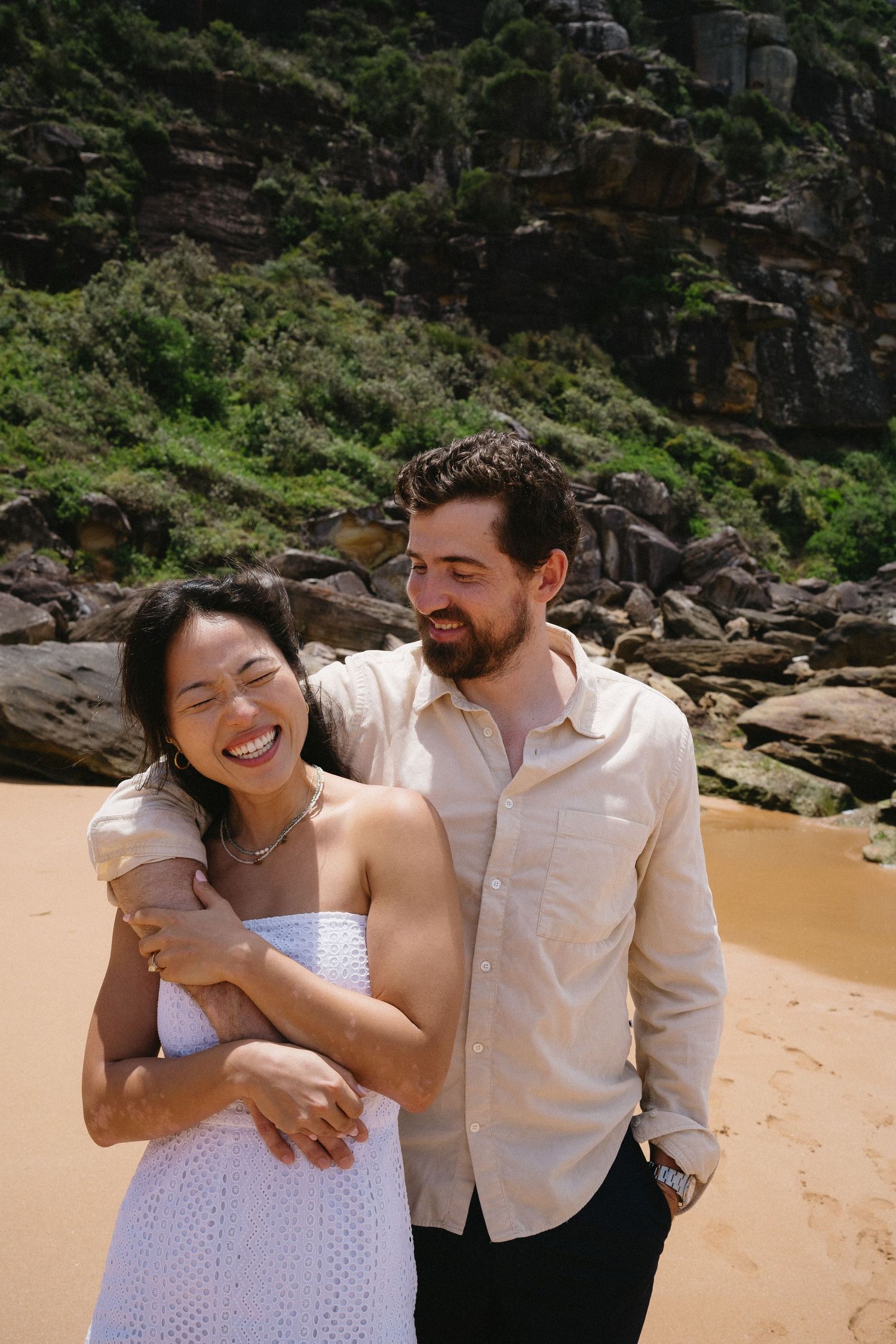 Happy couple laughing together on a beach with rugged cliffs and ocean waves behind them.