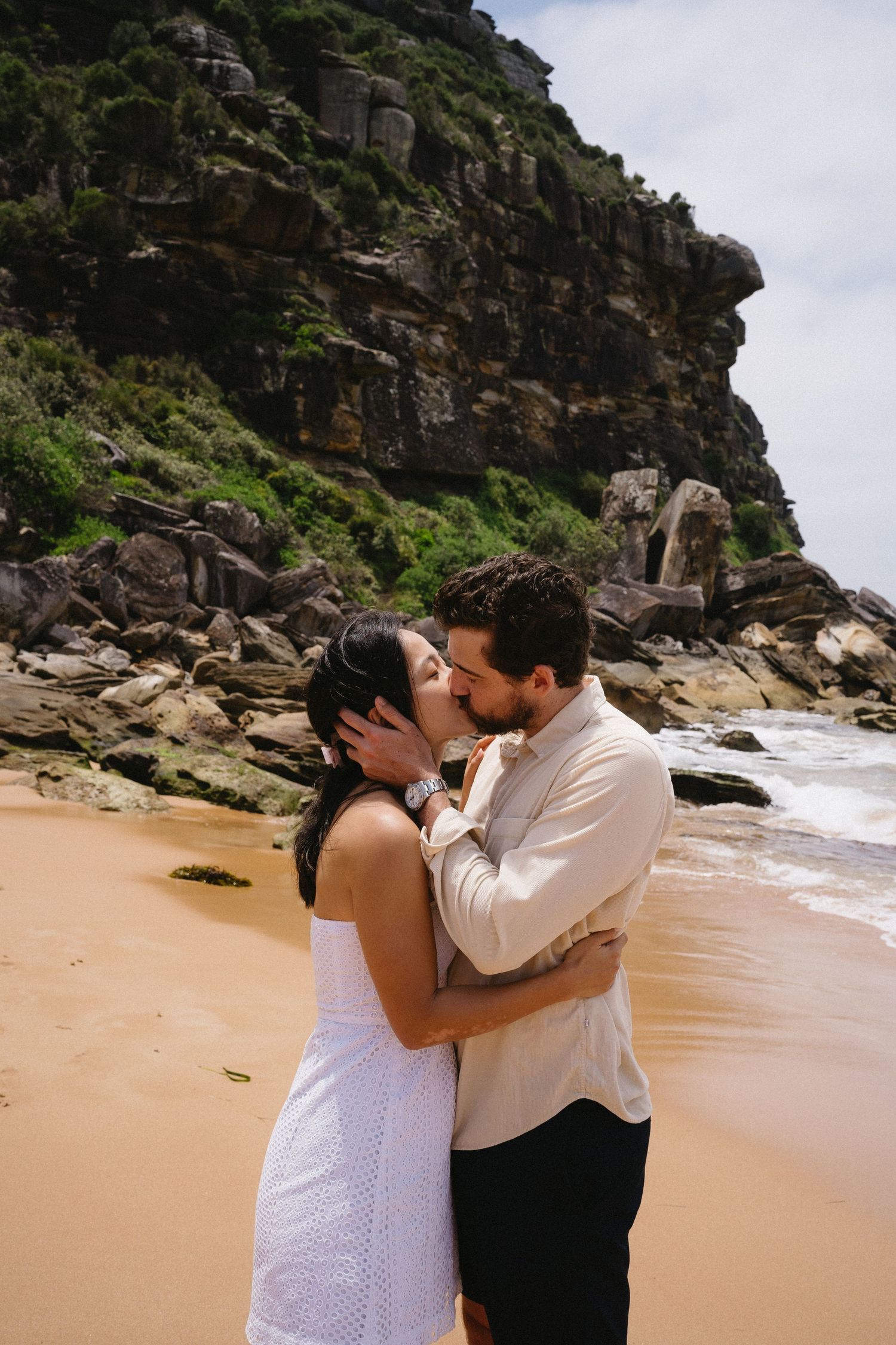 A couple shares a romantic kiss on a scenic beach with dramatic cliffs and rocky coastline in the background.