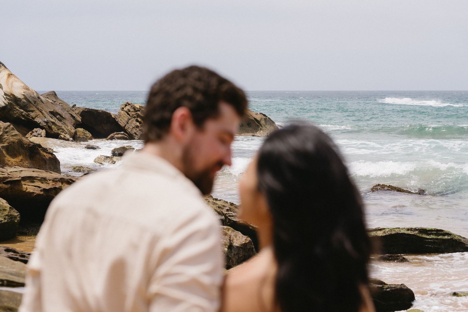 Intimate moment between couple on beach with crashing waves and coastal rocks.